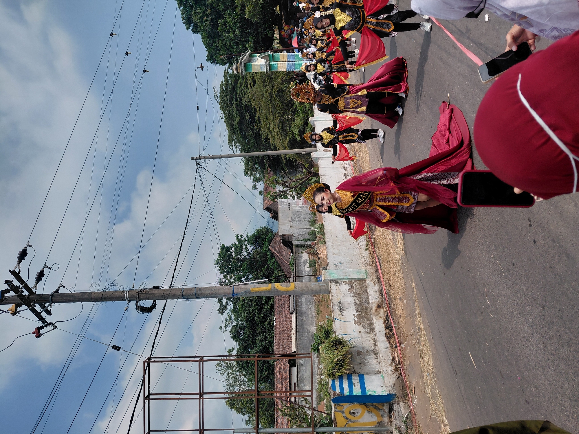 A group of young women in elaborate traditional costumes are participating in a parade on a paved street in Bimo, Indonesia. The central figure wears a crimson gown with gold embellishments and a golden headdress, holding a sash that reads "MIT MAIN". Other participants are dressed in black and gold attire with flowing red fabrics. They are walking in formation, holding hands or holding the fabric as part of their performance. The background shows a residential area with low-rise buildings and tiled roofs, interspersed with trees. A prominent utility pole with numerous wires is visible on the left side of the street. In the foreground, two individuals are partially visible, holding up smartphones, presumably to record or photograph the parade. The sky is partly cloudy, suggesting it is daytime. The overall scene depicts a cultural event or celebration taking place within a community.