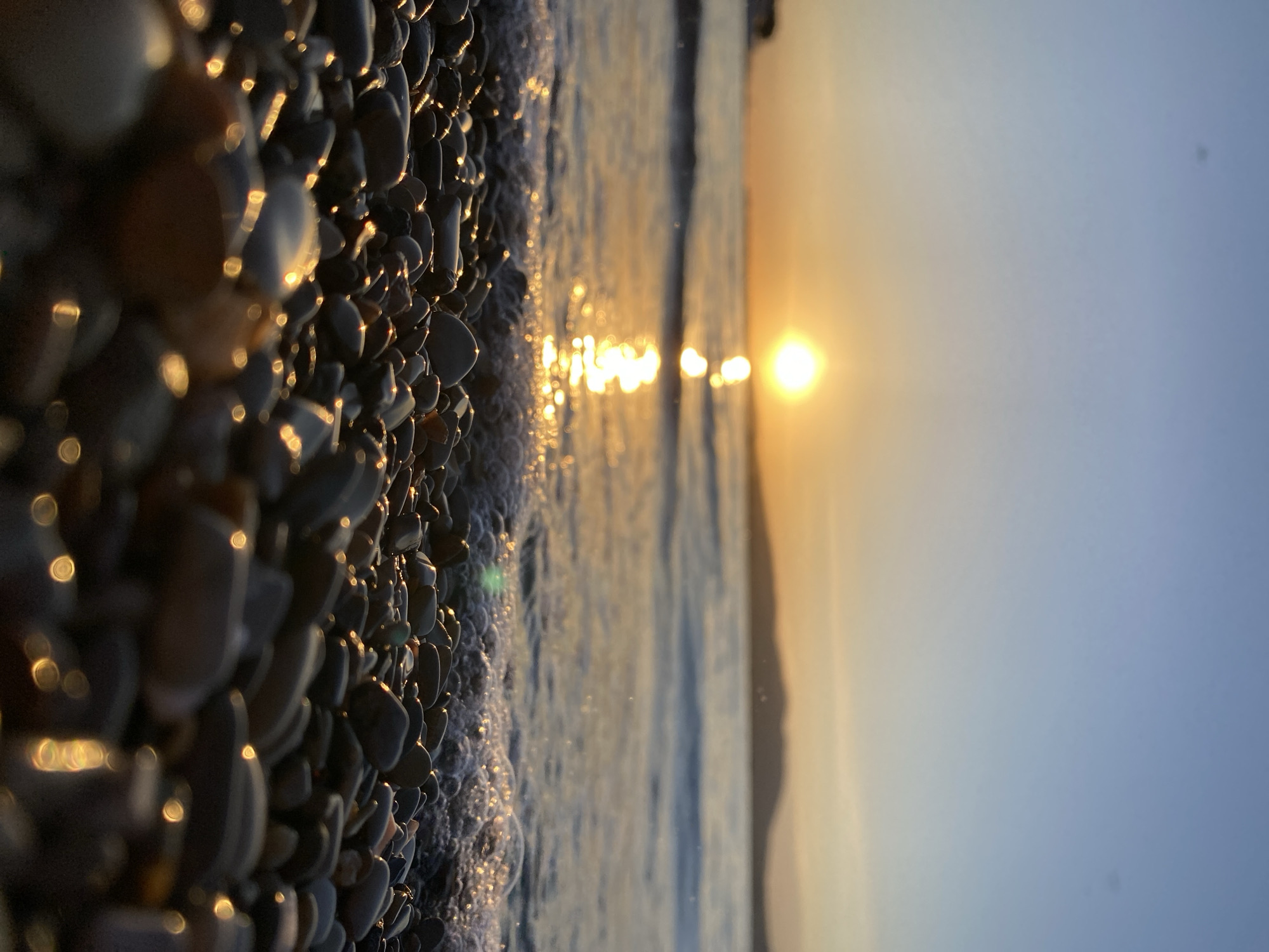 This image captures a serene close-up view of a pebble beach at the water's edge, likely during sunrise or sunset. The scene is set on a coastline, possibly in Gelendzhik, Russia, which is known for its pebble beaches.
In the foreground, numerous smooth, dark-colored pebbles cover the ground, wet from the sea. Many of these stones brilliantly reflect the golden light of the sun, creating bright, circular and elliptical bokeh highlights that add a shimmering quality to the texture of the beach.
Moving towards the midground, the edge of the water is visible, with gentle, white-foamed waves softly lapping against the pebbles. The water's surface reflects the sun's intense light, forming a shimmering path extending from the horizon.
The background is dominated by a bright, golden sun, positioned low on the horizon. It casts a warm, radiant glow across the water and into the sky. The sky transitions from a soft, warm orange-yellow near the sun to a clear, pale blue at the top of the frame. Faint, dark silhouettes of distant land, possibly hills or mountains, are discernible along the horizon line.
The image evokes a sense of peace and natural beauty, emphasizing the interplay of light, water, and the unique texture of the pebble beach. There are no people or discernible text visible in the frame.