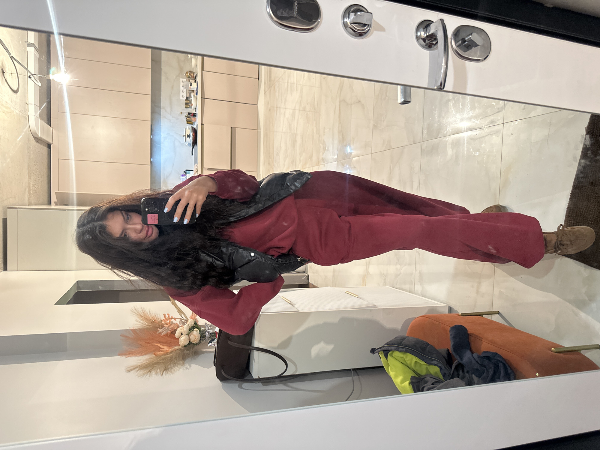 A woman is taking a selfie in a mirror. She is wearing a maroon jumpsuit and a black puffy vest. Her long dark hair is loose. She is holding a black smartphone with light blue nail polish on her fingers. The reflection shows a modern interior with light-colored cabinets, tile flooring, and a white console table. There's a bouquet of flowers next to the console. An orange ottoman and a jacket are visible in the lower right. The location may be an apartment or home in a city like Khimki, Russia, inferred from the general style of the interior.