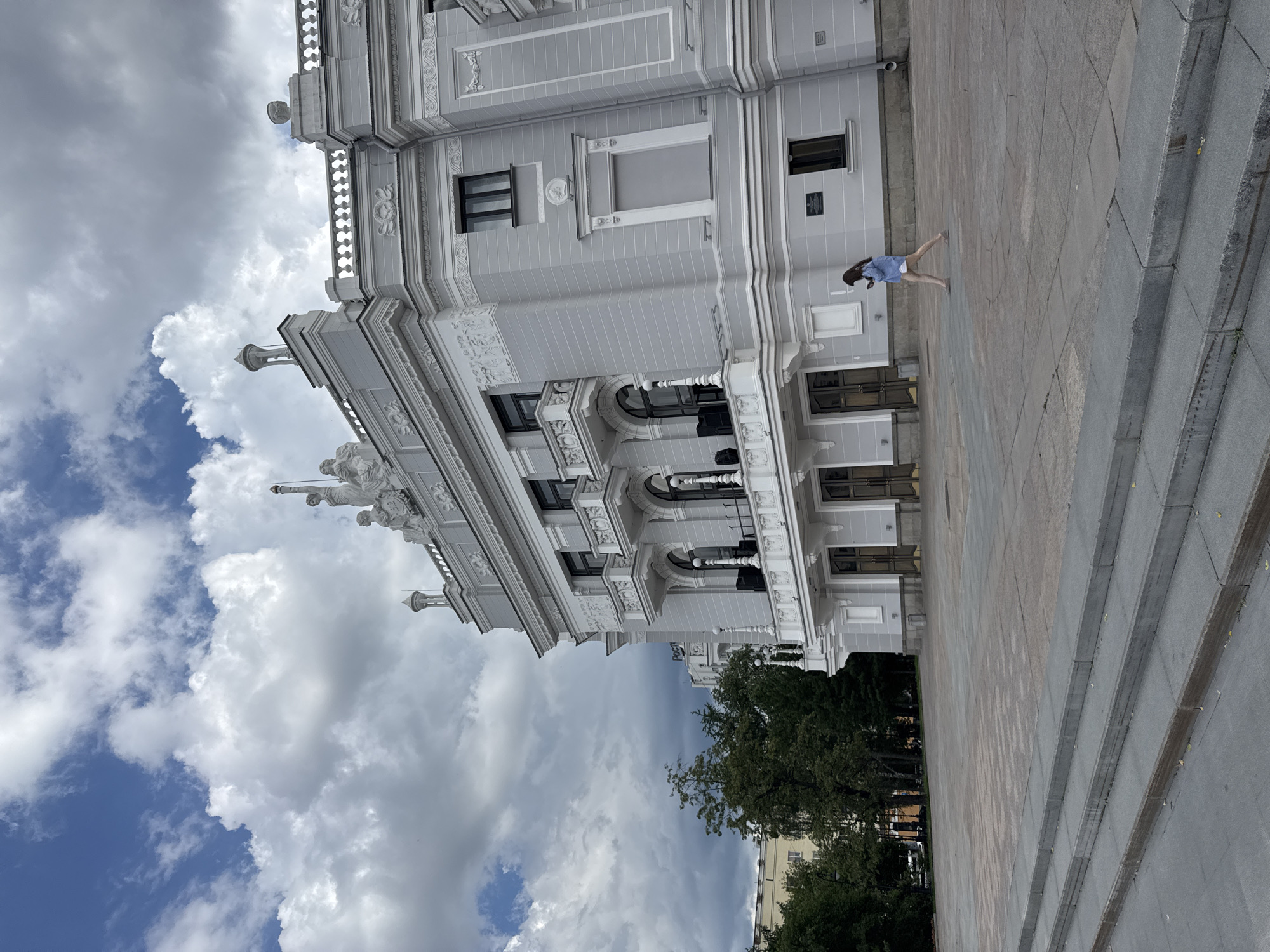 The image shows the Yekaterinburg Opera and Ballet Theatre, located in Yekaterinburg, Russia. A young woman in a blue shirt and white shorts is walking across the paved square in front of the building. The theatre is a grand, light gray building with ornate architectural details, including balconies, statues on the roofline, and decorative carvings. The sky above is filled with dramatic, cloudy formations. Trees are visible to the left of the theatre. A set of wide, concrete stairs leads up to the foreground of the square.