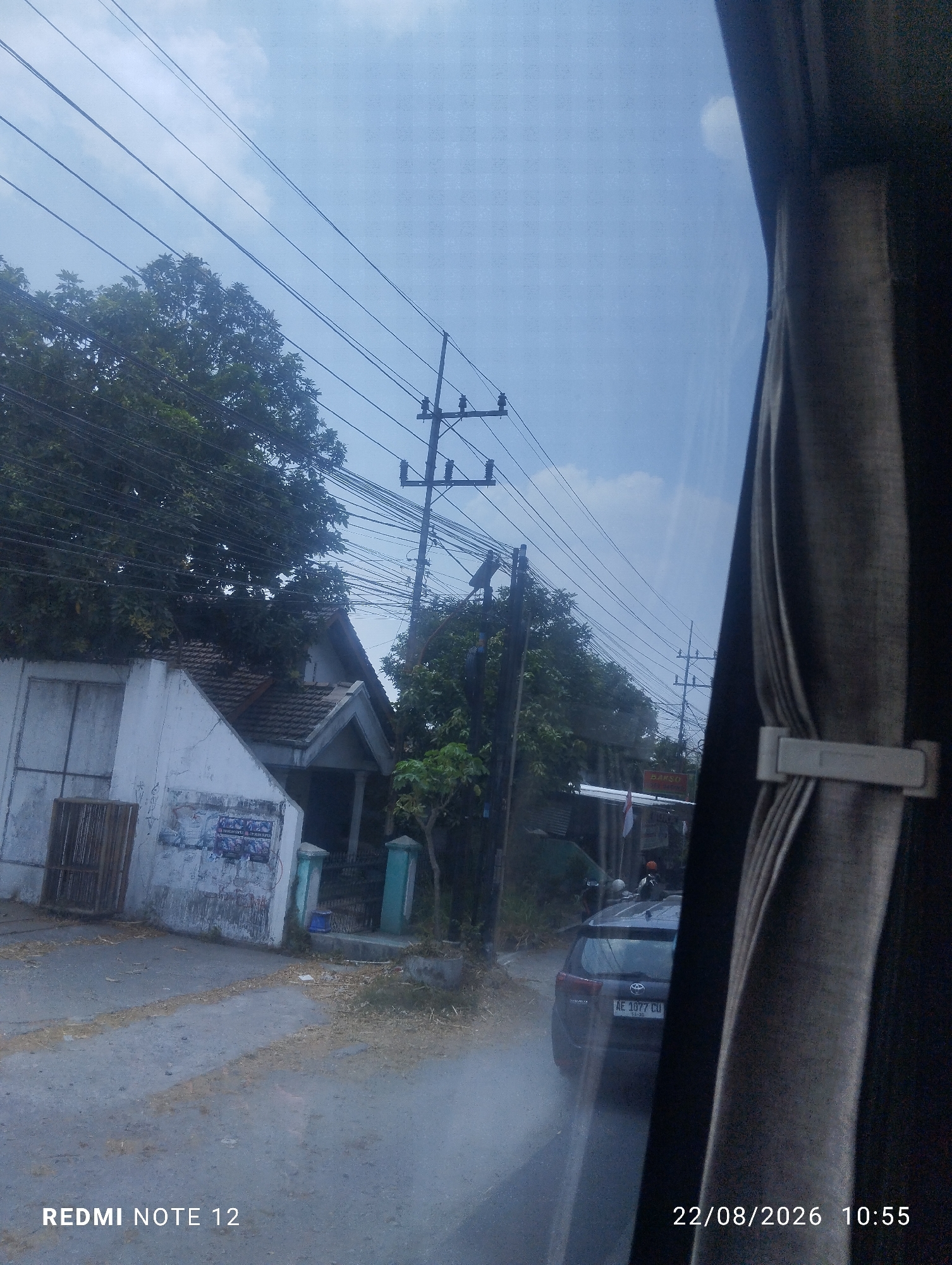 The image captures a street view in Kraton, Indonesia, with a focus on a residential area alongside a road. A dark-colored vehicle with license plate "AE 1077 CU" is parked on the right side of the road. In the background, a traditional Indonesian house with a tiled roof is visible, partially obscured by lush green trees and dense power lines that crisscross the sky. A white wall with posters adorns the front of a building on the left, with a wooden fence to its left. The sky is clear blue with scattered clouds. The lower right corner of the image displays a timestamp: "22/08/2026 10:55" and "REDMI NOTE 12".