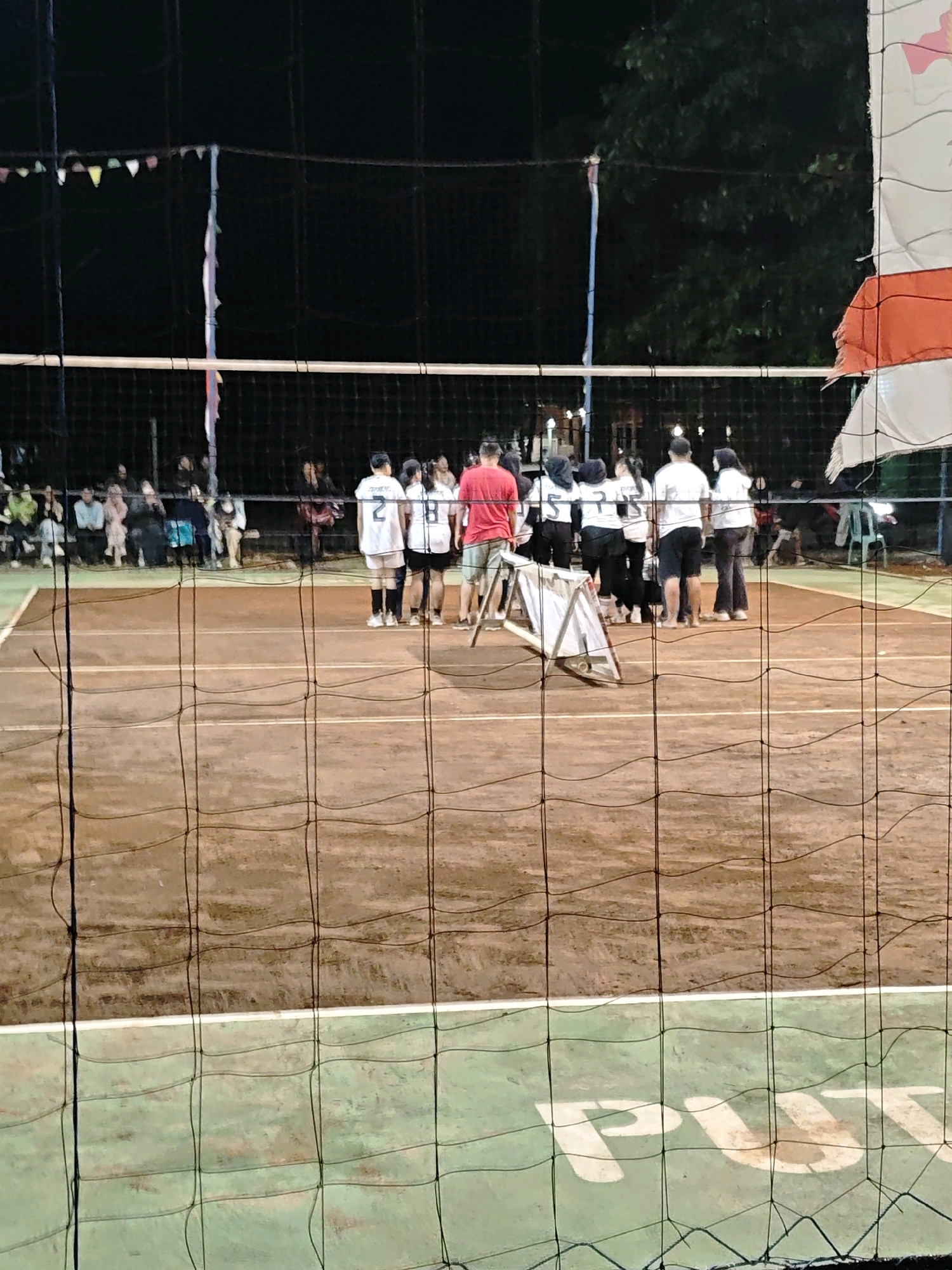 The image depicts a volleyball court at night in Pagu, Indonesia. A group of people, likely players and spectators, are gathered near the net. Several individuals in athletic attire are on the court, with one person in a red shirt standing prominently. The court itself appears to be made of clay, with a green outer perimeter marked with white lines. A net divides the court, and a white triangular structure is positioned near the center. In the background, trees and a dark sky are visible, suggesting an outdoor setting. The presence of string lights and spectators further indicates a public event. The overall scene captures a moment of a sports gathering, possibly a game or practice, under artificial lighting.