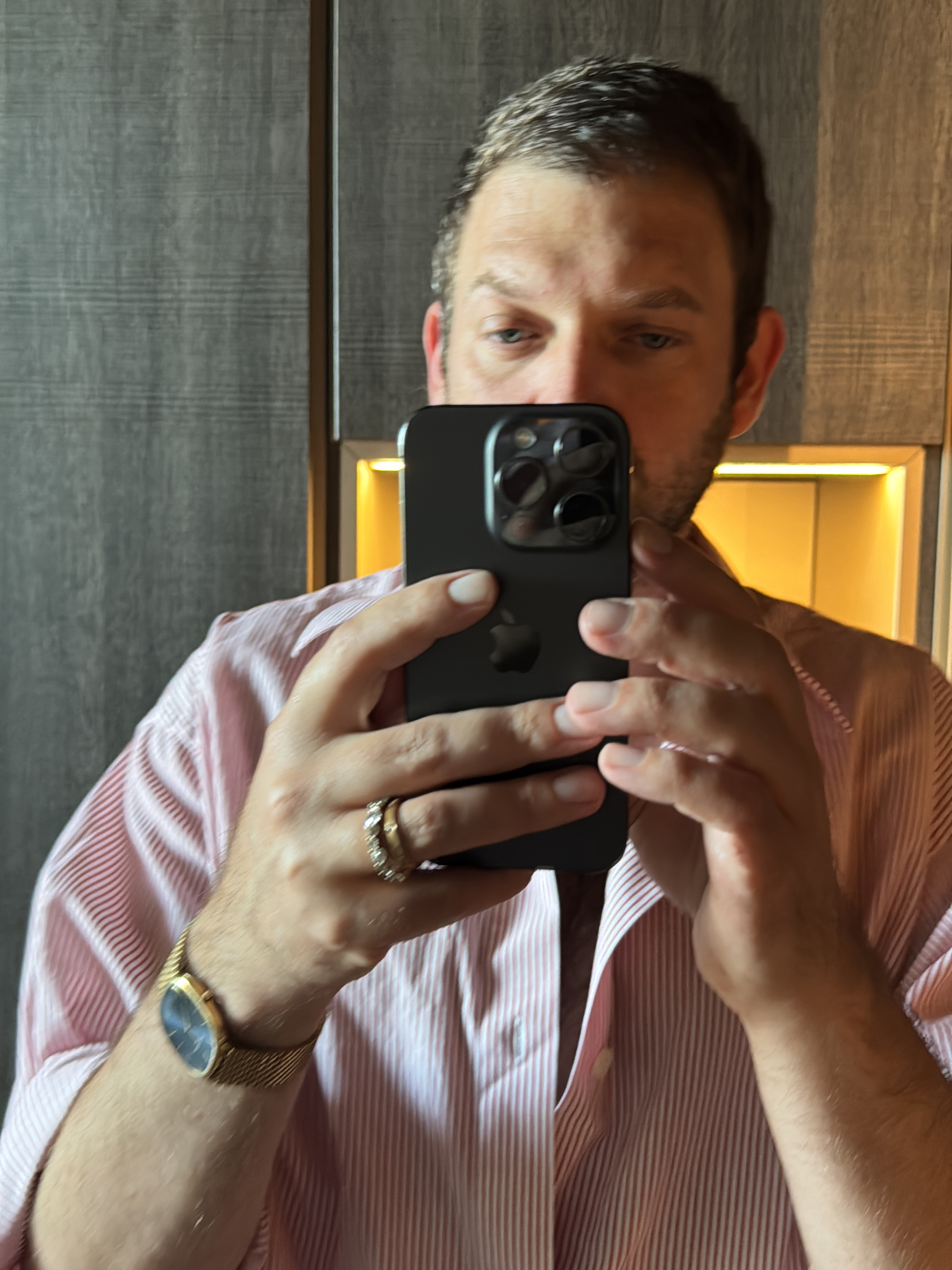 The image is a close-up, slightly distorted selfie taken by a man holding a dark-colored smartphone with multiple cameras. He is wearing a pink and white striped collared shirt, partially unbuttoned. On his left hand, he wears two rings, one appearing to be a wedding band with small stones. A gold watch with a mesh band is visible on his left wrist. The background appears to be an interior setting, possibly a hotel room or apartment, with textured grey paneling on the left and illuminated golden paneling on the right. The lighting suggests an indoor environment. The man is looking at the phone's screen, indicating he is taking the photo. The scene does not provide any direct information about the city of Milan, Italy.