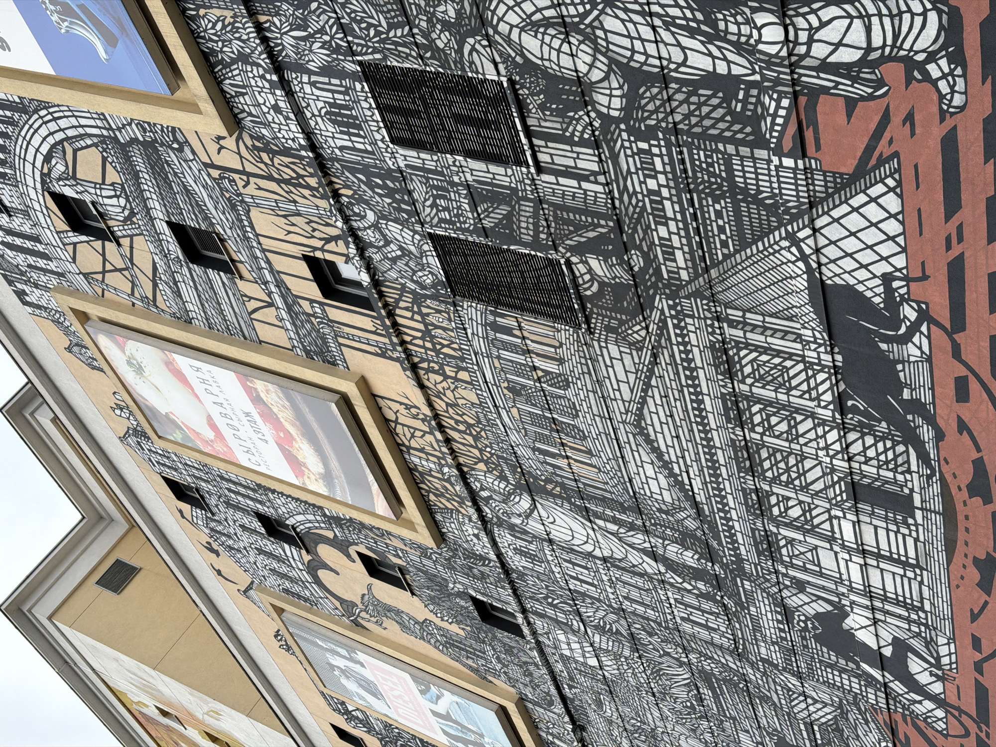 The image depicts a low-angle view of a building facade in Moscow, Russia. The building is adorned with a large-scale, black-and-white graphic mural depicting architectural elements and abstract patterns. Several framed advertising posters are affixed to the building, interspersed with the mural. The mural incorporates the building's architectural features like windows and ventilation grilles into its design. A section of the mural has a salmon-colored accent. The sky is overcast.