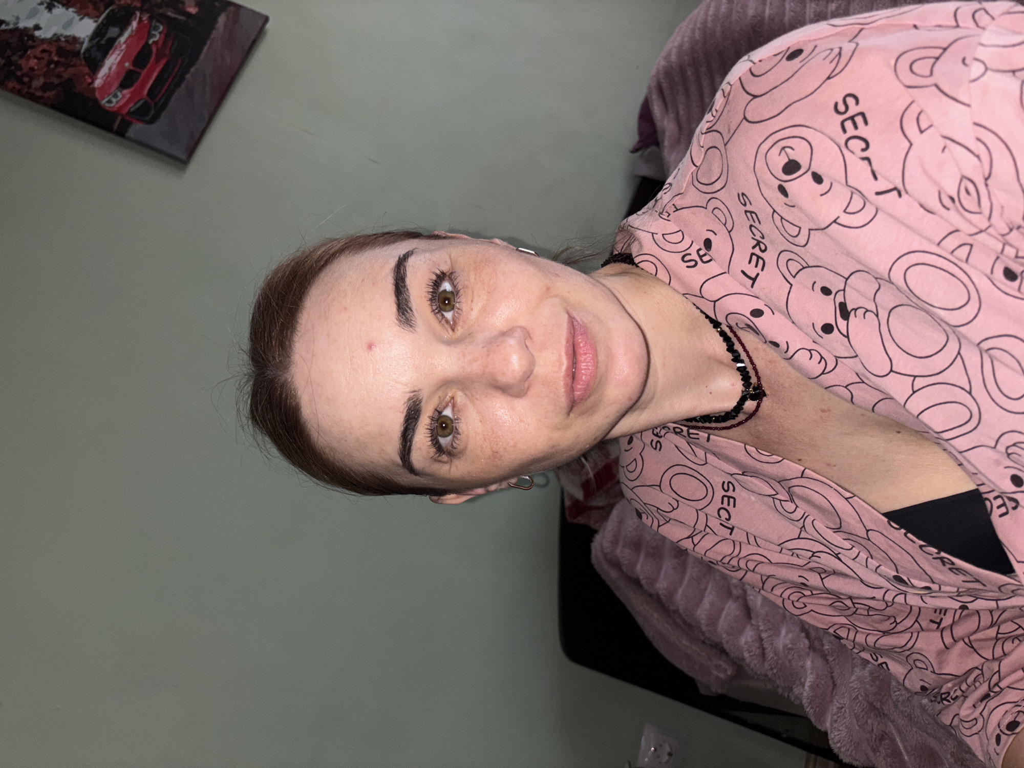 A woman with light skin, brown hair pulled back, and brown eyes is shown from the chest up, looking directly at the camera. She is wearing a pink robe with a pattern of teddy bears and the word "SECRET" repeated throughout. She has a black necklace with red beads and a dark pendant. She is also wearing small silver hoop earrings. A pink fuzzy blanket is visible behind her to the left, and a black chair is partially visible. To the right of the woman, on a light green wall, is a framed picture of a red car. The overall environment appears to be indoors, possibly a bedroom.