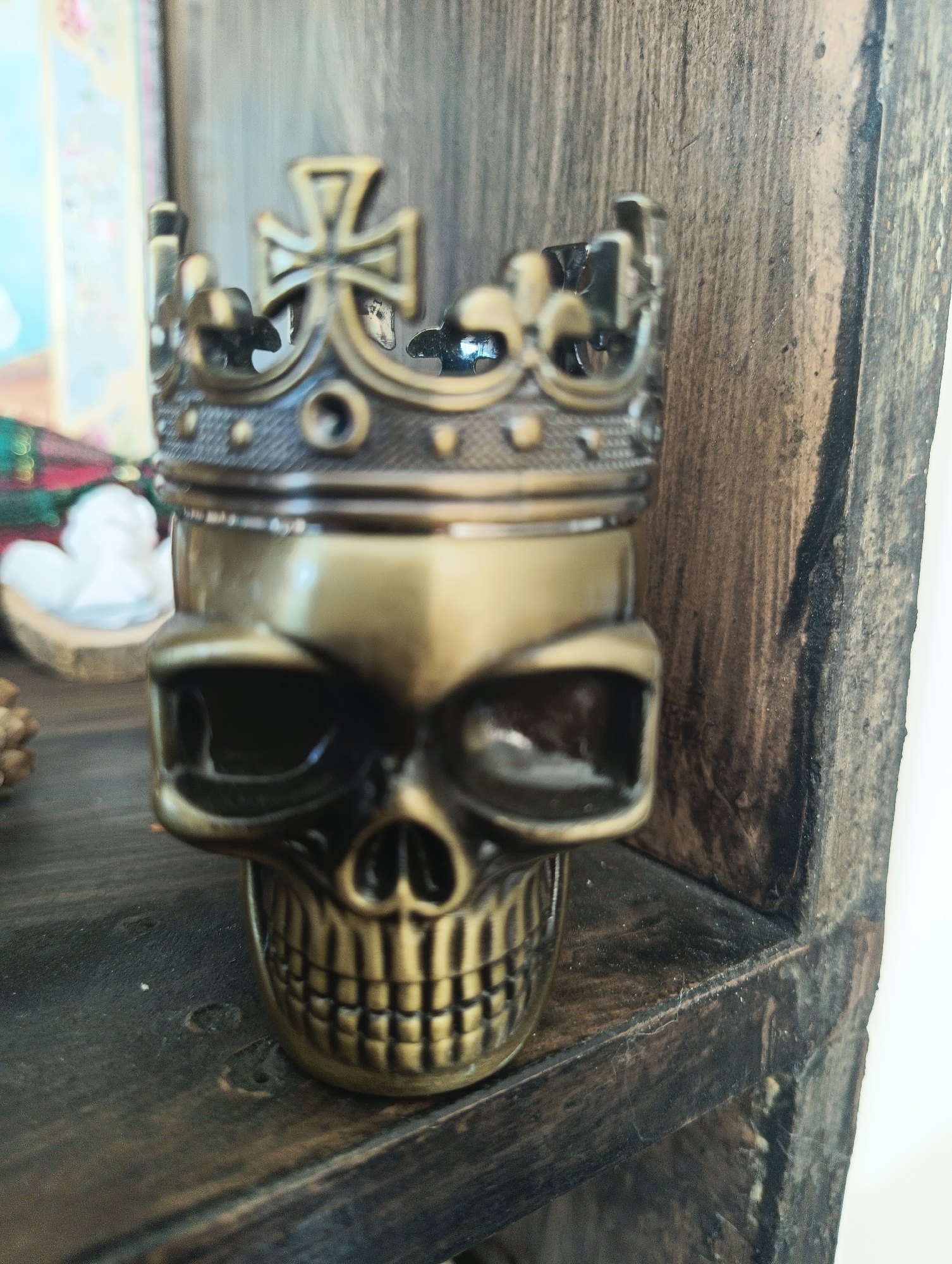 The image shows a close-up of a brass-colored skull figurine wearing a crown. The figurine is placed on a dark wooden shelf. In the background, out of focus, there are some decorative items, including what appears to be a white angel figurine and some greenery. The overall aesthetic is dark and somewhat gothic. There is no information in the image to suggest a location in the city of Karnobat, Bulgaria.