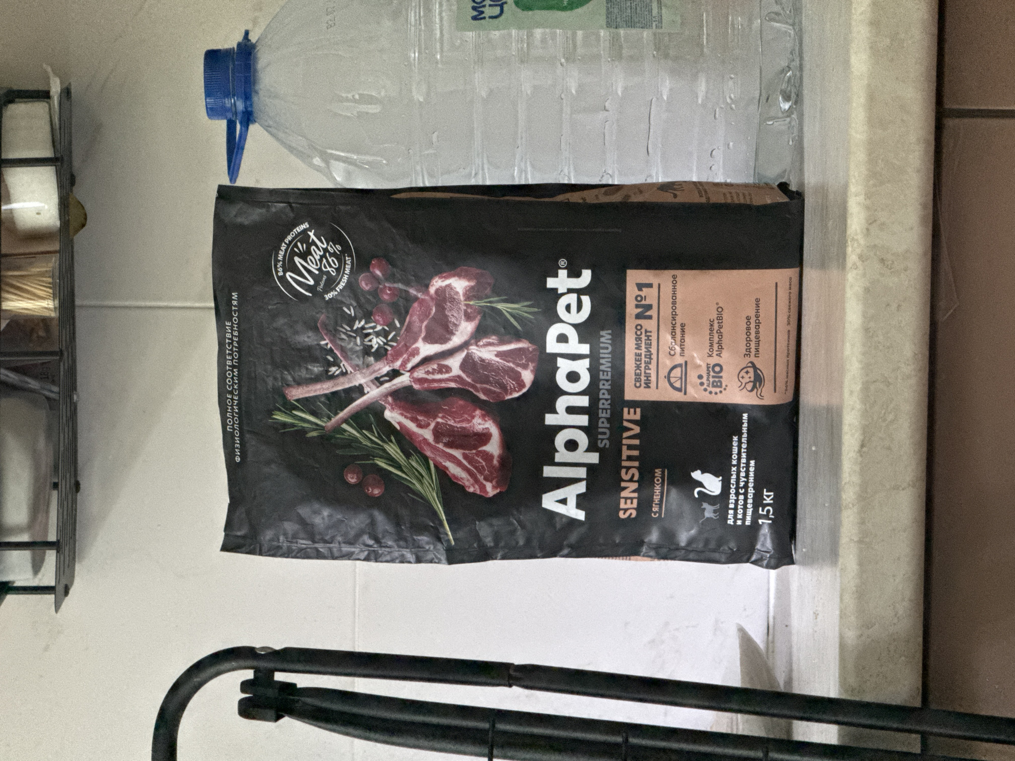 This is a medium-shot, eye-level photograph of a bag of AlphaPet cat food. The bag is black with images of lamb chops, cranberries, and rosemary. The text on the bag is in Russian and English, indicating that it is "SENSITIVE" food with "86% MEAT PROTEINS" and "30% FRESH MEAT." The bag is placed on a countertop next to a large, clear plastic bottle of water with a blue cap. In the background, a metal shelf with toothpicks is visible. The environment appears to be a domestic kitchen, with tiled walls and a countertop. The lighting is natural, coming from a window or overhead source, and casts a slight shadow on the countertop. The image does not provide any specific information about its location within the city of Saratov, Russia.