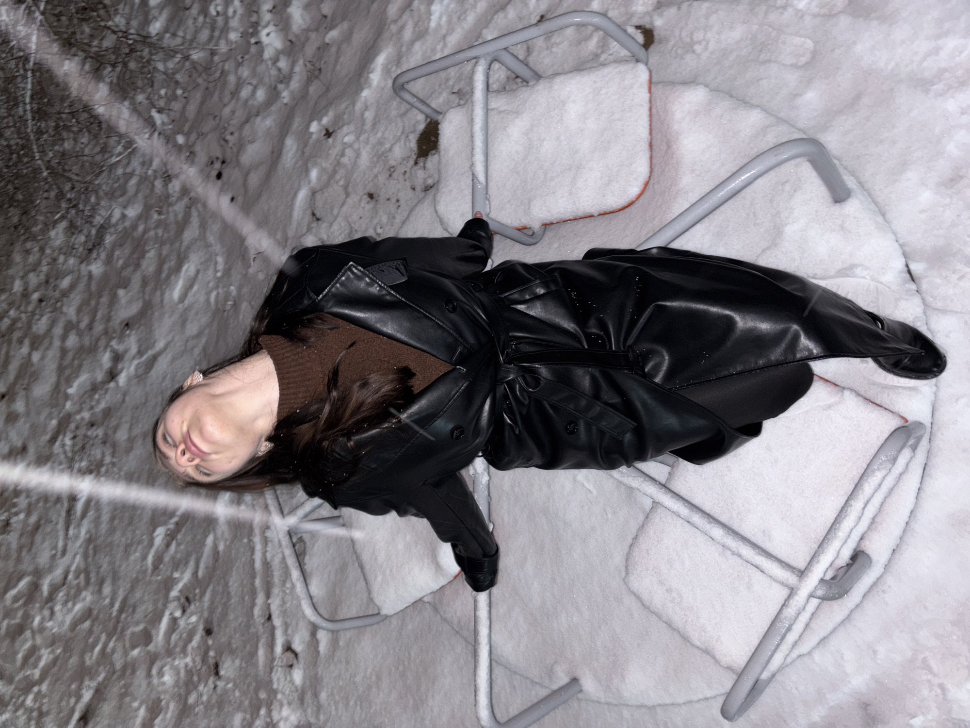 The image displays a young woman lying on a snow-covered child's merry-go-round at night in Perm, Russia. The scene is dominated by a thick layer of fresh, white snow covering the ground and the grey metal frame of the playground equipment.
The woman is positioned centrally, her body stretched out on the merry-go-round's seat and the surrounding snow. She is dressed in a dark brown high-neck top and a long, belted black faux leather coat. Her long, dark brown hair spreads out behind her head. Her face is turned slightly upwards, with eyes mostly closed and a faint, relaxed smile, conveying a calm and somewhat playful demeanor.
The time of day is nighttime, indicated by the dark background and the prominent artificial lighting, which casts a bright streak across the snow to the left and illuminates her face and coat. The weather is clearly cold and snowy, with small white specks, likely snowflakes, visible on her coat and in the air. Bare tree branches can be seen on the left side of the frame, confirming the winter setting. No visible text is present in the image.