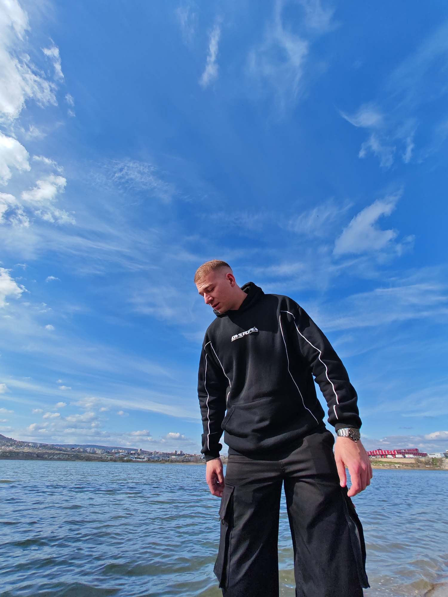 A man stands in water, wearing a black hoodie with white trim and black cargo pants. The background features a blue sky with wispy clouds, a body of water, and a shoreline with buildings. The man appears to be standing in Lake Zawierzbie near the town of Divdyadovo in Shumen, Bulgaria.
