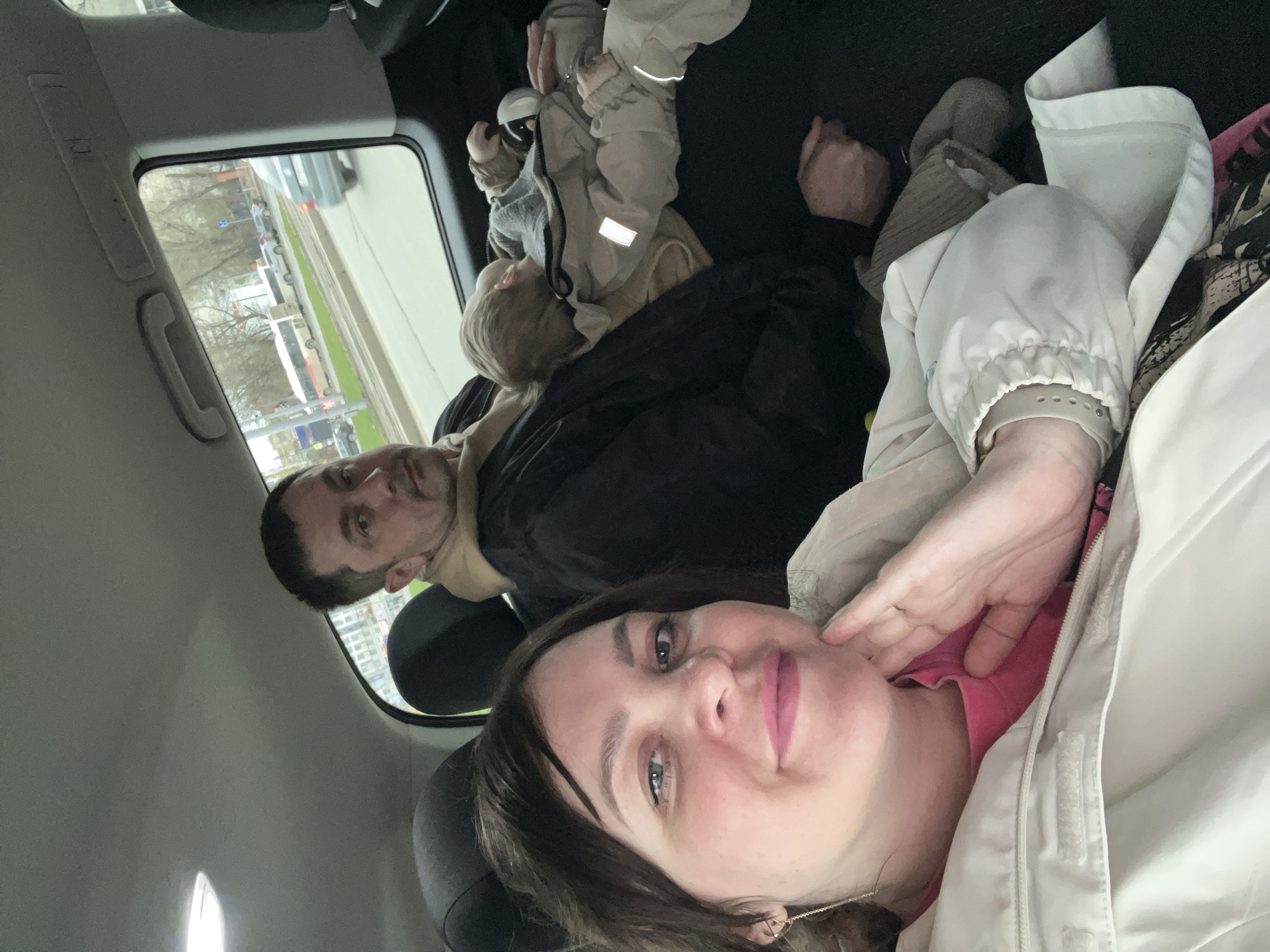 The photograph shows four individuals inside a vehicle, likely a car. A woman is positioned in the foreground, appearing to be taking a selfie. Behind her to the left, a man is visible looking towards the camera. Behind the man are two children. The view out the car window reveals a street scene in Perm, Russia.