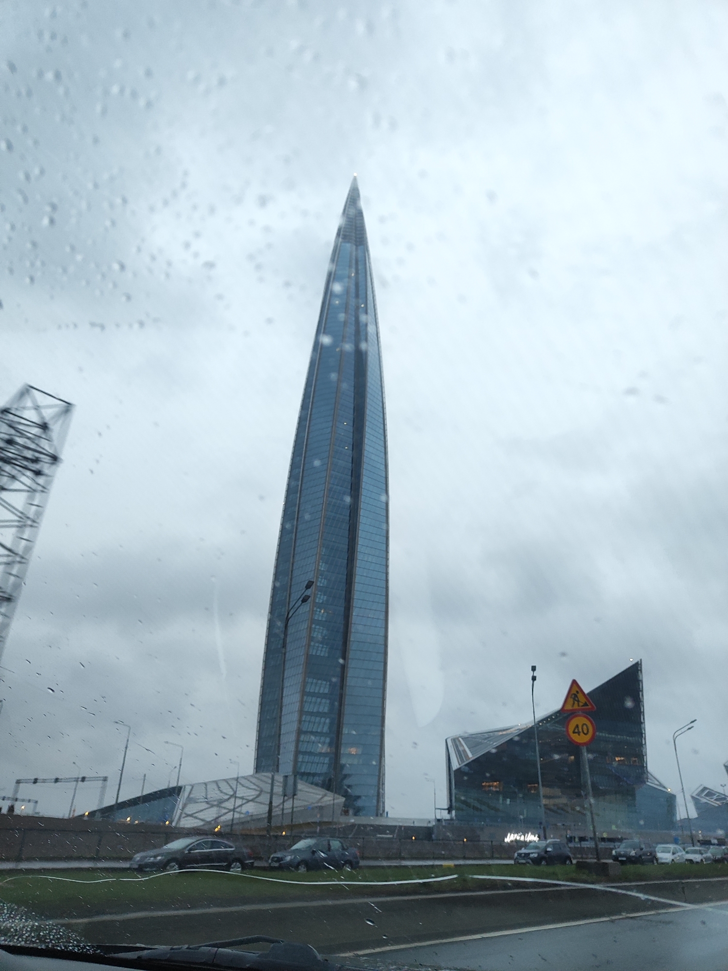 The image shows a tall, glass-clad skyscraper in an urban setting, likely taken through a vehicle windshield covered in raindrops. To the left, there is a metal structure, possibly a transmission tower or part of an unfinished building. Adjacent to the skyscraper is a lower, modern-looking building with an angular design, featuring glass facades. A road runs in front of the buildings with parked cars. A traffic sign with a "40" speed limit is visible. The sky is overcast, suggesting a rainy or gloomy day. Based on the architecture, this appears to be the Lakhta Center in Saint Petersburg, Russia.