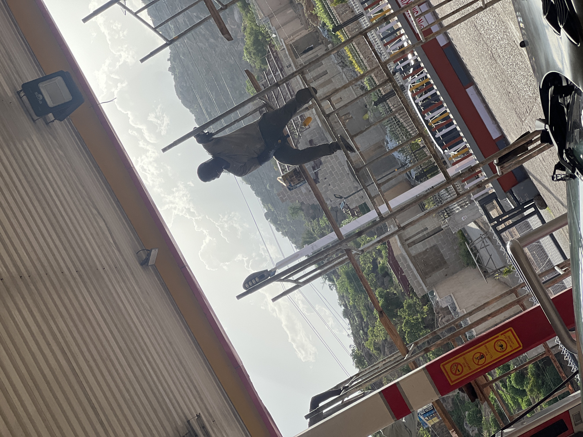 The image depicts an outdoor scene in Gura Top, Nigeria. A man wearing a dark shirt and pants is climbing on a scaffolding structure. The scaffolding appears to be constructed from metal poles and is erected in front of what seems to be a building under construction. The background shows a hillside with green vegetation and a cloudy sky. A sign with pictograms indicating "No Smoking" and "No Telephones" is visible on the scaffolding. Part of a vehicle, possibly a motorcycle, is in the extreme right of the frame. A modern outdoor light fixture is attached to the side of the building.