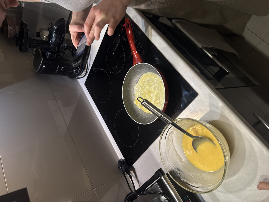 A person takes a photo with their phone of an electric stovetop with a frying pan on it. A thin pancake is cooking in the pan. A glass bowl containing batter with a spoon rests near the stovetop. A black juicer sits on the counter beside the stovetop. The countertop and floor are light-colored tile. A kitchen cabinet is in the background.
