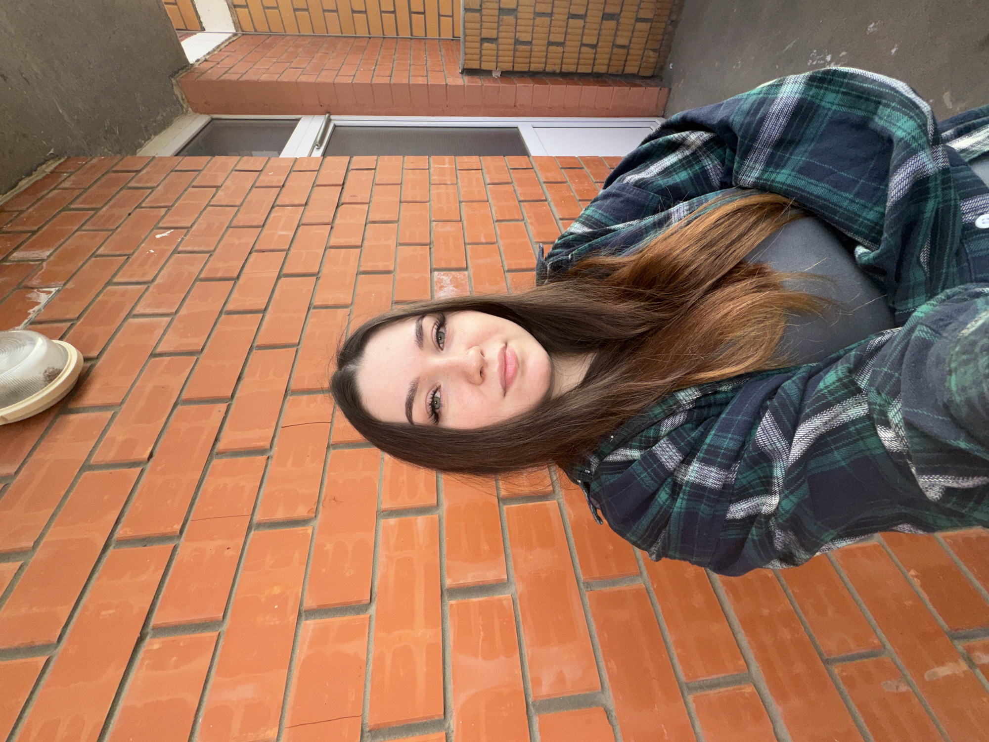 A young woman with long brown hair stands against a brick wall in what appears to be an outdoor urban setting. She is wearing a dark green and navy plaid shirt over a grey top. The brick wall behind her is orange and appears to be part of a building. To her right, there is a white door and a portion of another brick wall. The lighting suggests it is daytime. This scene is consistent with an exterior location in the city of Perm, Russia, possibly a residential building or commercial establishment with exposed brick architecture.