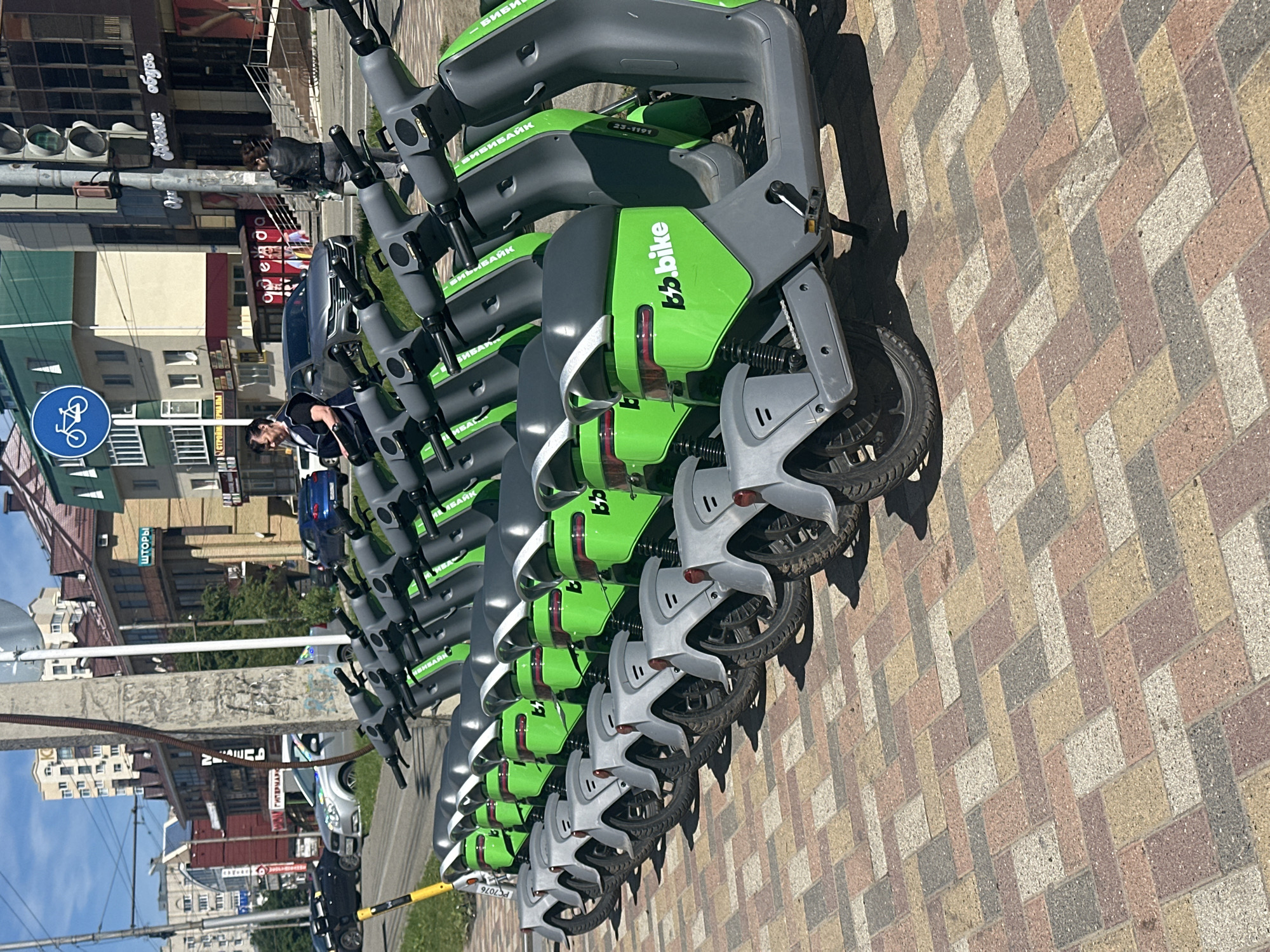 This image captures a bright, sunny urban scene in Stavropol, Russia, dominated by a long row of parked electric scooters. The scooters, part of the "b.bike" (БИБИБАЙК) sharing service, are uniformly grey with lime green accents and are neatly lined up on a multi-colored paved sidewalk, which features a herringbone pattern. Their strong, dark shadows indicate direct sunlight, suggesting it is midday. Several scooters display identification numbers like "23-1191" and "PC7076".
In the background, a busy city street unfolds with various multi-story buildings. Storefronts are visible, displaying Cyrillic text such as "ГИПЕРМАРКЕТ" (Hypermarket, partially visible), "ШТОРЫ" (Curtains), and "Несие обувь" (likely a shoe store). Cars are present on the street, both parked and in motion. A few people can be seen in the background; one individual in dark clothing stands near the line of scooters, while another is further back by a dark car.
Urban infrastructure elements include traffic lights and a prominent blue circular sign with a white bicycle icon, indicating a bicycle path or zone. The scene depicts a modern city environment utilizing shared electric mobility solutions.