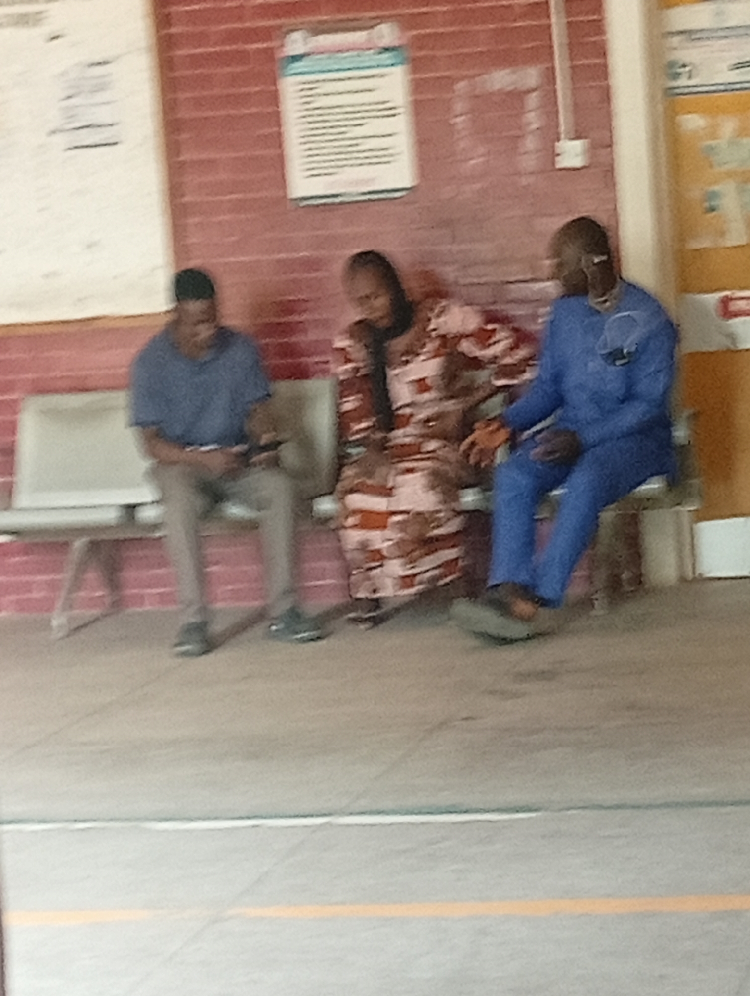 Three individuals are seated on a bench in front of a brick wall, with a white and a yellow poster displayed on the wall. The person on the left is wearing a blue t-shirt and grey pants, looking down at their phone. The person in the middle is wearing a patterned dress and is partially obscured. The person on the right is wearing a blue tracksuit and is wearing headphones around their neck. The floor is concrete with yellow and white lines. The overall scene appears to be a waiting area in a public facility, potentially in Gombe, Nigeria, given the context.