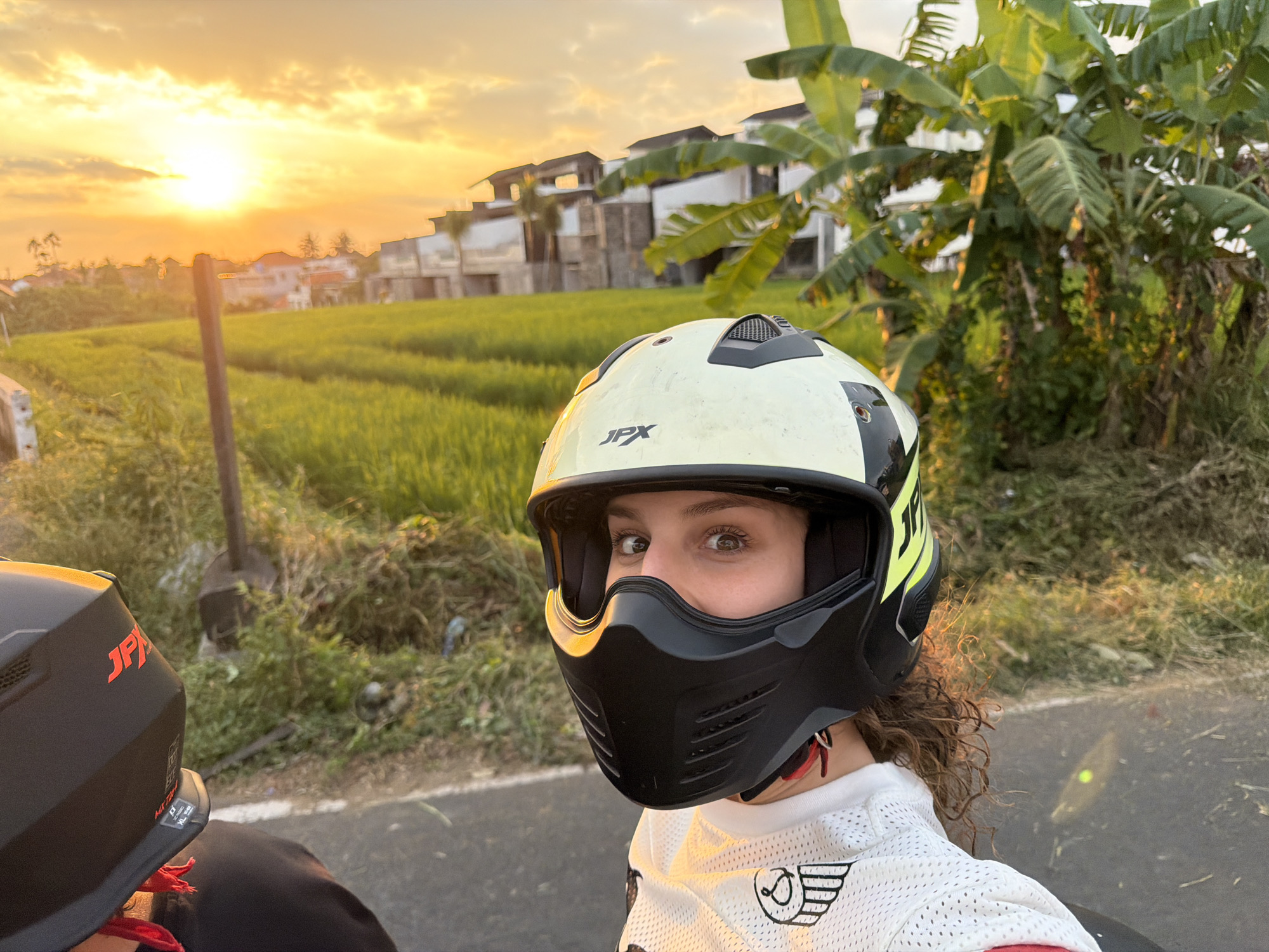 The image shows a person wearing a black and white full-face motorcycle helmet, with the front of the helmet facing the camera. The person is wearing a white jersey with black graphics. Their eyes are visible through the helmet's visor. The scene is outdoors, with a golden sunset casting a warm glow. To the right of the person, there are lush green rice fields and some buildings, suggesting a rural or semi-rural setting. Banana trees are also visible in the foreground. The image is oriented vertically. The location is specified as North Kuta, Indonesia.