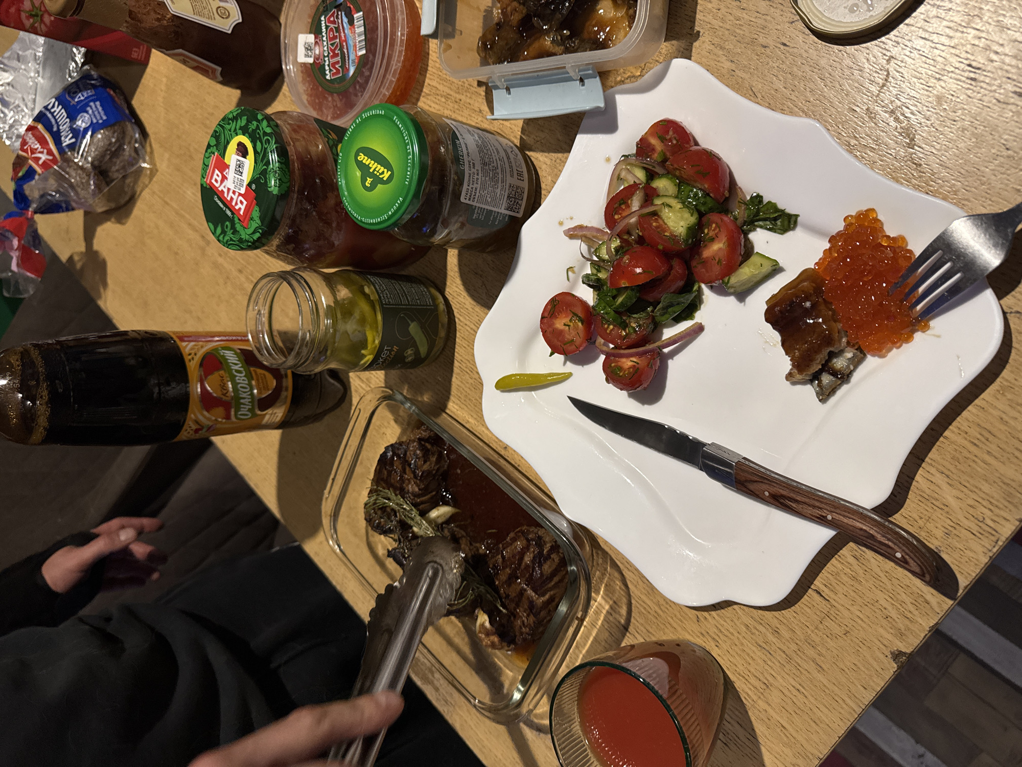 The image shows a table setting with various food items and drinks. On the table, there are jars of pickles, a container of what appears to be red caviar, a container of stewed food, and a loaf of bread in a plastic bag. A white plate contains a salad of tomatoes, cucumbers, and red onions, as well as a serving of fish topped with red caviar, which is being held by a fork. A knife is also on the plate. A glass container holds cooked meat with sauce, and a pair of tongs is being used to handle the meat. A glass of red liquid, possibly tomato juice or sauce, is also present. A bottle of Очаковский (Ochakovsky) kvass is also visible.
Based on the labels on the food items, the image includes items from Russia, such as the "ВАНЯ" brand pickles and "ДАРЫ САХАЛИНА" (Gifts of Sakhalin) caviar, as well as Kühne pickles.
Ptichnoye is a location only in Ukraine and there is no clear association with the scene.