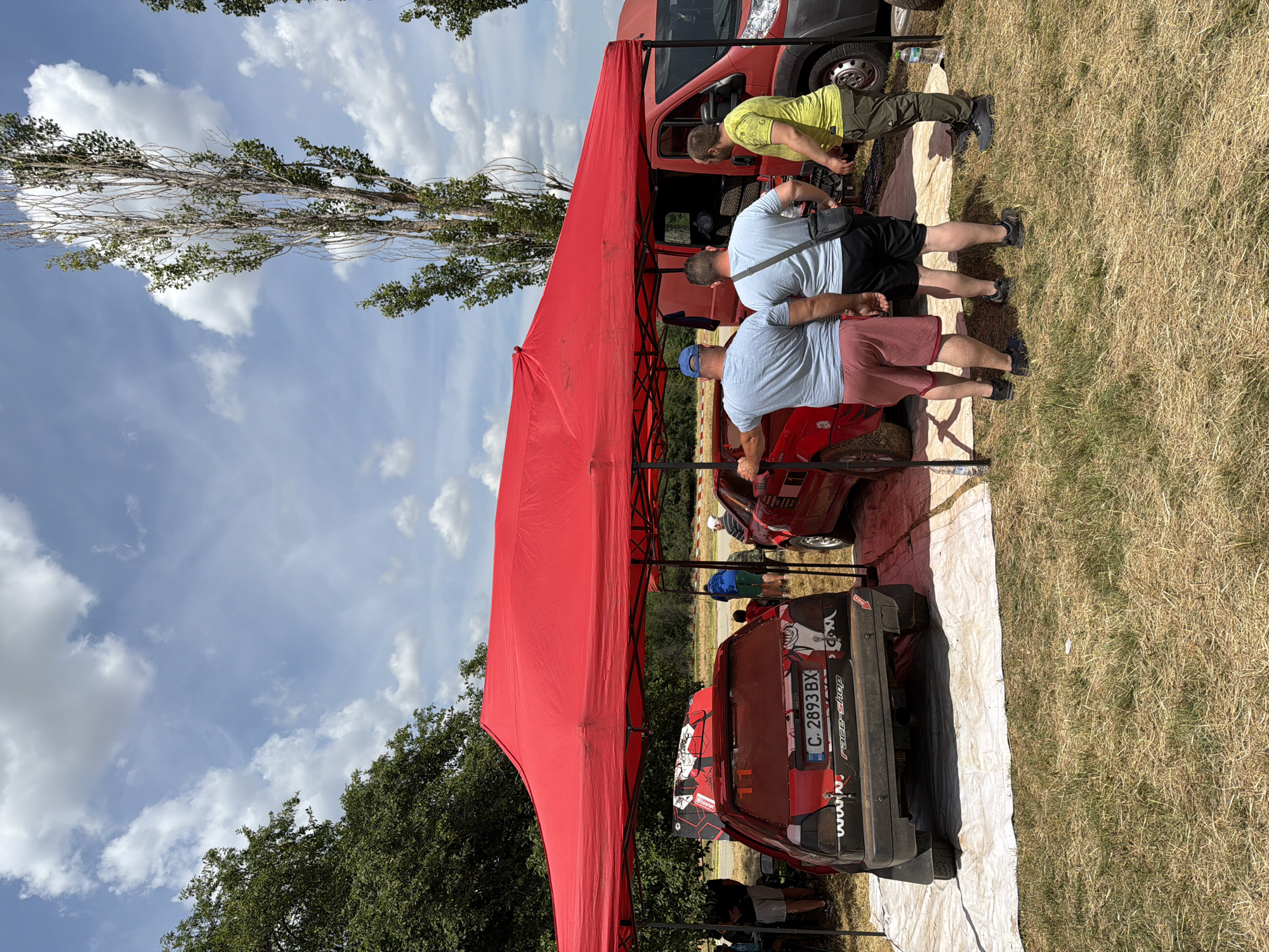 In Bahovitsa, Bulgaria, under a bright, partly cloudy sky, a group of individuals are gathered near a large red tent. The tent provides shade for two rally cars, one black and one red, both bearing racing decals and stickers. The black car has a license plate that reads "C 2893 BX". A red van is parked to the right of the tent. Three men in casual attire are standing together, facing the red car. One man wears a blue cap and red shorts, another a grey shirt and black shorts, and the third a yellow t-shirt and green pants. The ground is covered with dry grass. The scene appears to be outdoors, possibly at a race event or a car show.