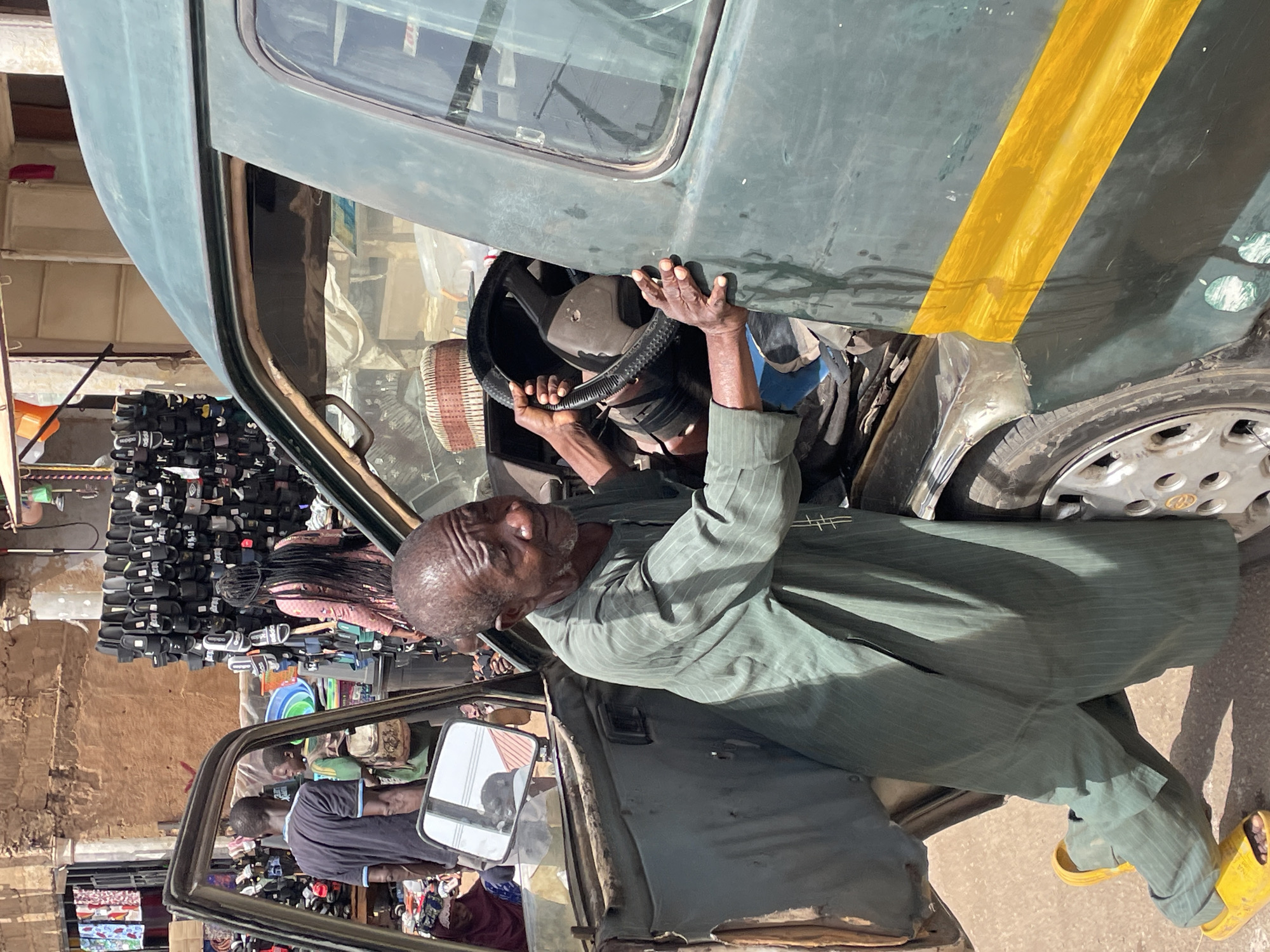 A medium shot of an elderly man seated in a green vehicle with a yellow stripe, holding the steering wheel. He is wearing a green striped suit and yellow sandals. The vehicle is parked on a street in Bukuru, Nigeria, where numerous shoes are displayed for sale. Other people are visible near the open door on the driver's side.