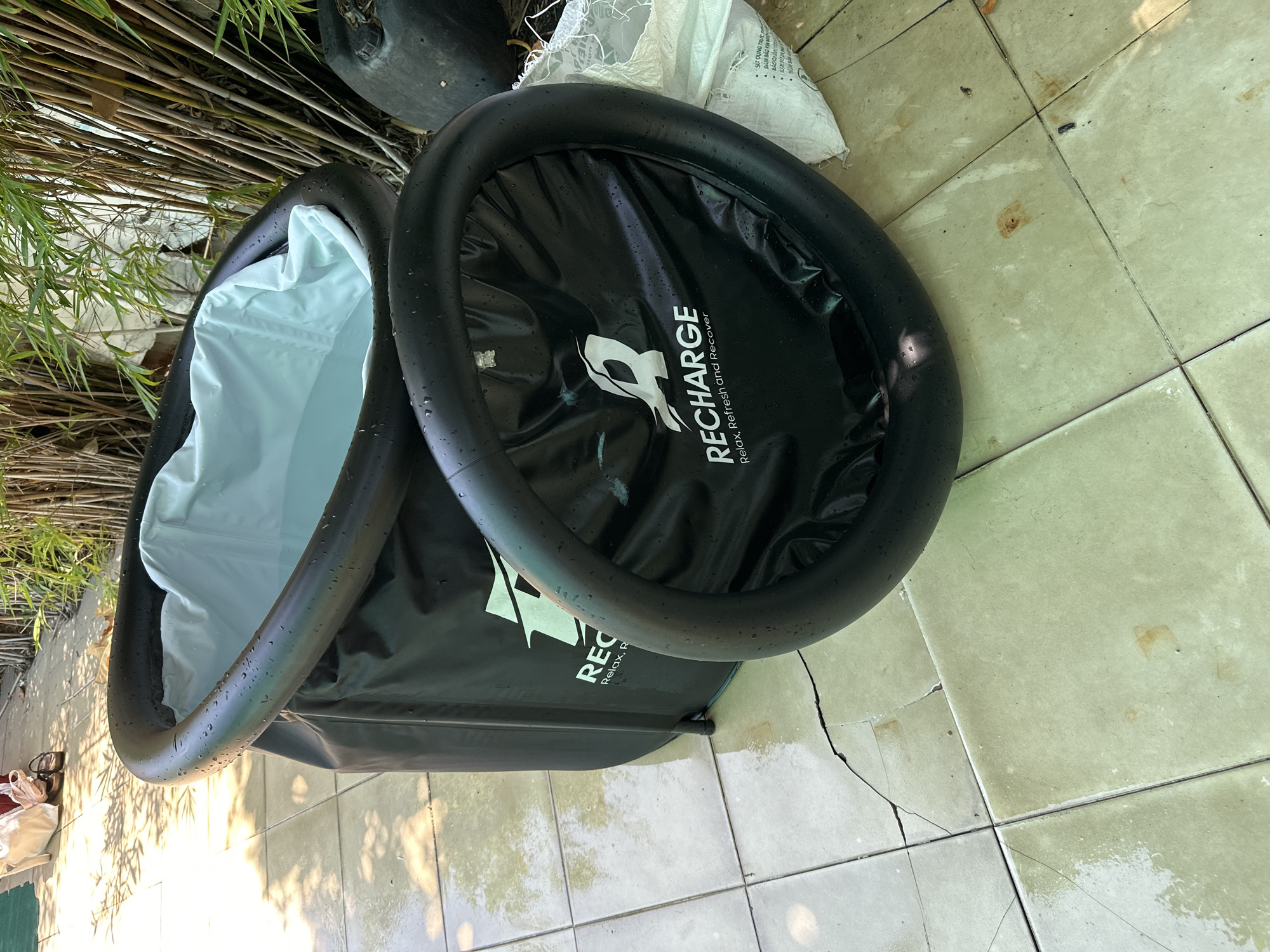 The image shows an inflatable black tub filled with water, resting on a tiled outdoor surface. The tub has a white inner lining and a thick black inflatable rim. The lid of the tub, also black with an inflatable rim, is lying next to it. Both the tub and the lid feature a white logo and the text "RECHARGE Relax, Refresh and Recover". The scene appears to be in an outdoor area with dried bamboo stalks in the background and dappled sunlight casting shadows on the ground. A dark plastic container is visible to the right of the tub. The tiled ground shows signs of moisture and some cracking. The overall setting suggests a residential or recreational outdoor space, possibly in Nha Trang, Vietnam, given the presence of bamboo commonly found in tropical regions and the style of the tiling. There are no people directly visible interacting with the tub.