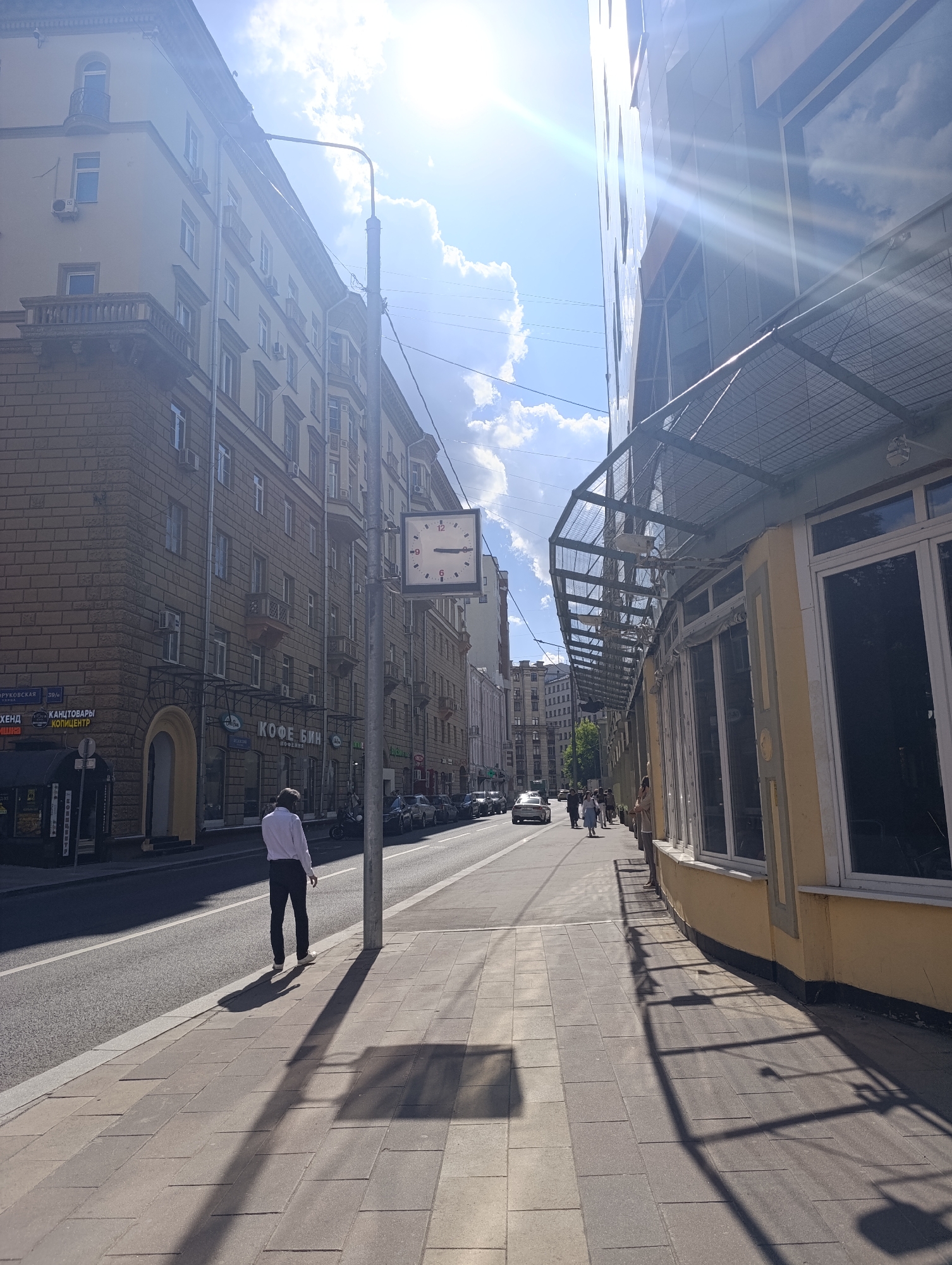 This bright, sunny photograph depicts a bustling street scene in Moscow, Russia, likely during late morning or early afternoon given the high sun and long shadows. The view looks down a wide city street flanked by multi-story buildings and populated by pedestrians and vehicles.
On the left, a traditional-style building with a beige facade features numerous windows, balconies, and air conditioning units. Its lower portion is faced with a textured, brick-like material. Ground-floor businesses display signs in Cyrillic, including "КОФЕ БИН" (Coffee Bean), "МЕБЕЛЬ" (Furniture), "канцтовары" (stationery), and "хоббицентр" (hobby center). A street sign on this building indicates "ВОРОНЦОВСКАЯ 39/4". Parked cars line the curb, and a black kiosk is visible near the corner.
A tall street lamp pole stands on the left sidewalk, centrally located, with a square-faced clock attached, indicating approximately 10:10. A man in a white shirt and dark pants walks away from the camera on this sidewalk, his long shadow stretching out.
The building on the right has a lighter, possibly yellow, facade on its ground floor, featuring large, reflective windows that mirror the blue sky and clouds. A prominent metal mesh awning extends over these windows. Several pedestrians are visible walking further down this sidewalk.
The sky is clear blue with scattered white clouds, and intense sunlight causes significant lens flare in the upper left of the image. Long, defined shadows are cast by the street lamp, pedestrians, and the building's awning onto the paved sidewalks. Overhead wires are visible against the bright sky. A white car and other vehicles are present on the street, suggesting active traffic.