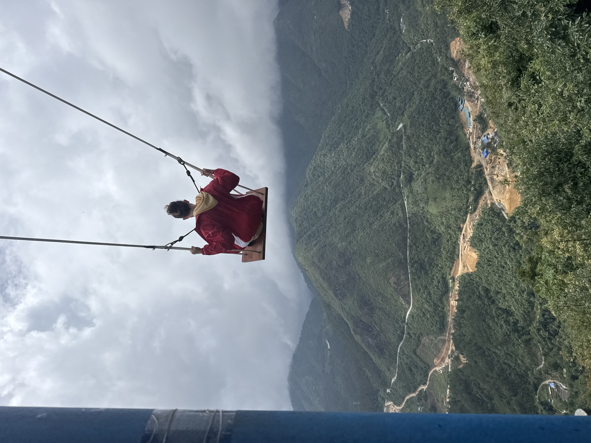 A person in a red jacket and brown shorts sits on a swing over a lush, mountainous valley. The swing is suspended by two ropes attached to a horizontal pole. The person is facing away from the camera. The valley below is green with dense vegetation, and a winding road can be seen. The sky is overcast with gray clouds. The location appears to be in a mountainous area near Binh Lu, Vietnam. A large blue metal bar is partially visible at the bottom of the image, likely part of a structure. A construction or industrial area is visible in the valley below.