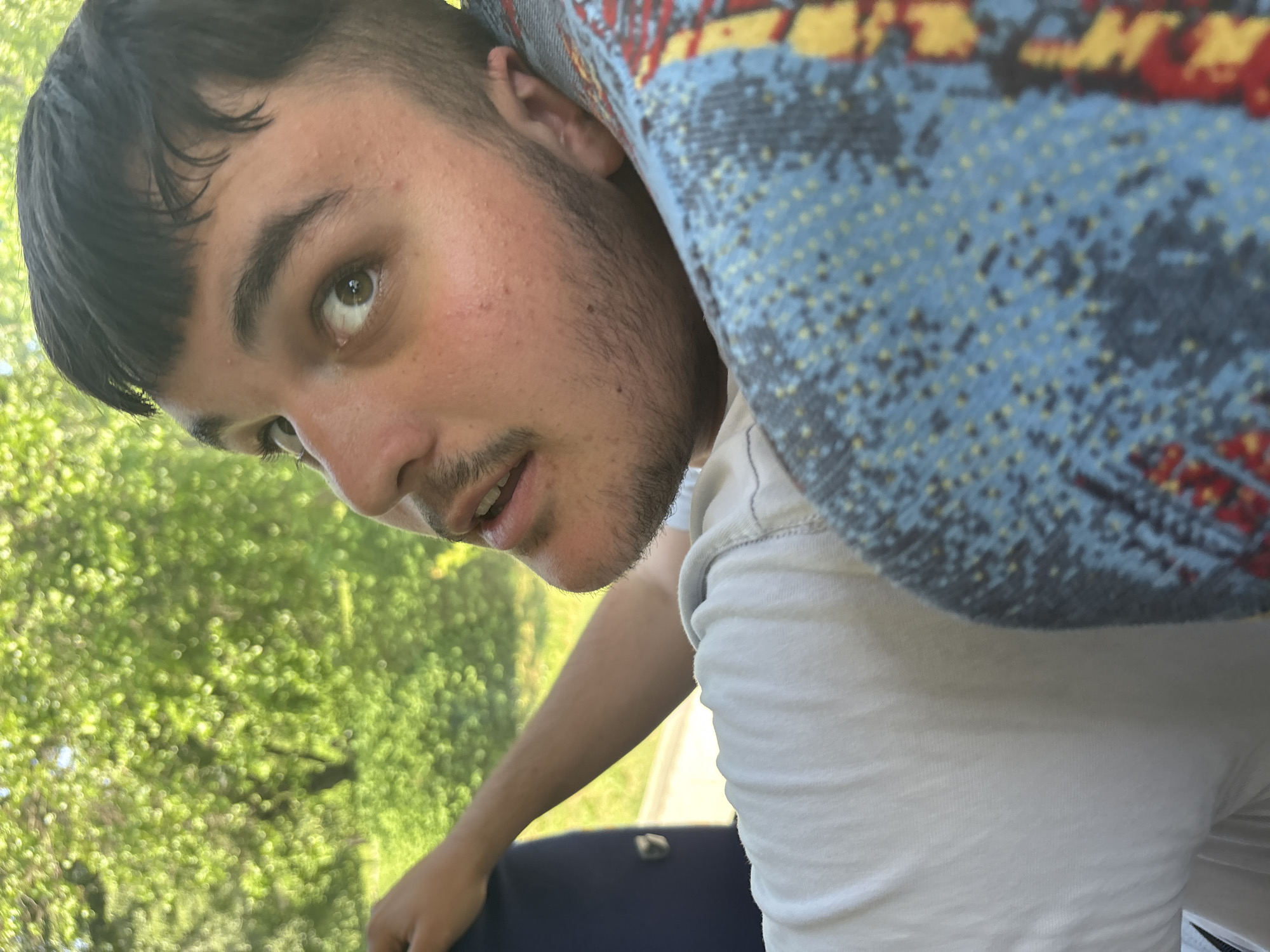 A young man with dark hair and light stubble is depicted in a close-up, angled shot. He is looking upwards and slightly to the left, with a curious or inquisitive expression. His mouth is slightly open, and he has light brown eyes. He is wearing a plain white t-shirt.
The man appears to be leaning back in a seat, possibly in a vehicle, as evidenced by a patterned headrest or seat fabric in the upper right of the frame. This fabric features an abstract design with blue as the primary color, accented with yellow, red, and darker blue or black shapes. Part of a dark blue seat is also visible in the lower left.
In the background, visible through what appears to be a window, is bright green, out-of-focus foliage, suggesting trees or bushes. The overall lighting is bright and natural, indicating it is daytime and likely sunny. No visible text is present in the image.
