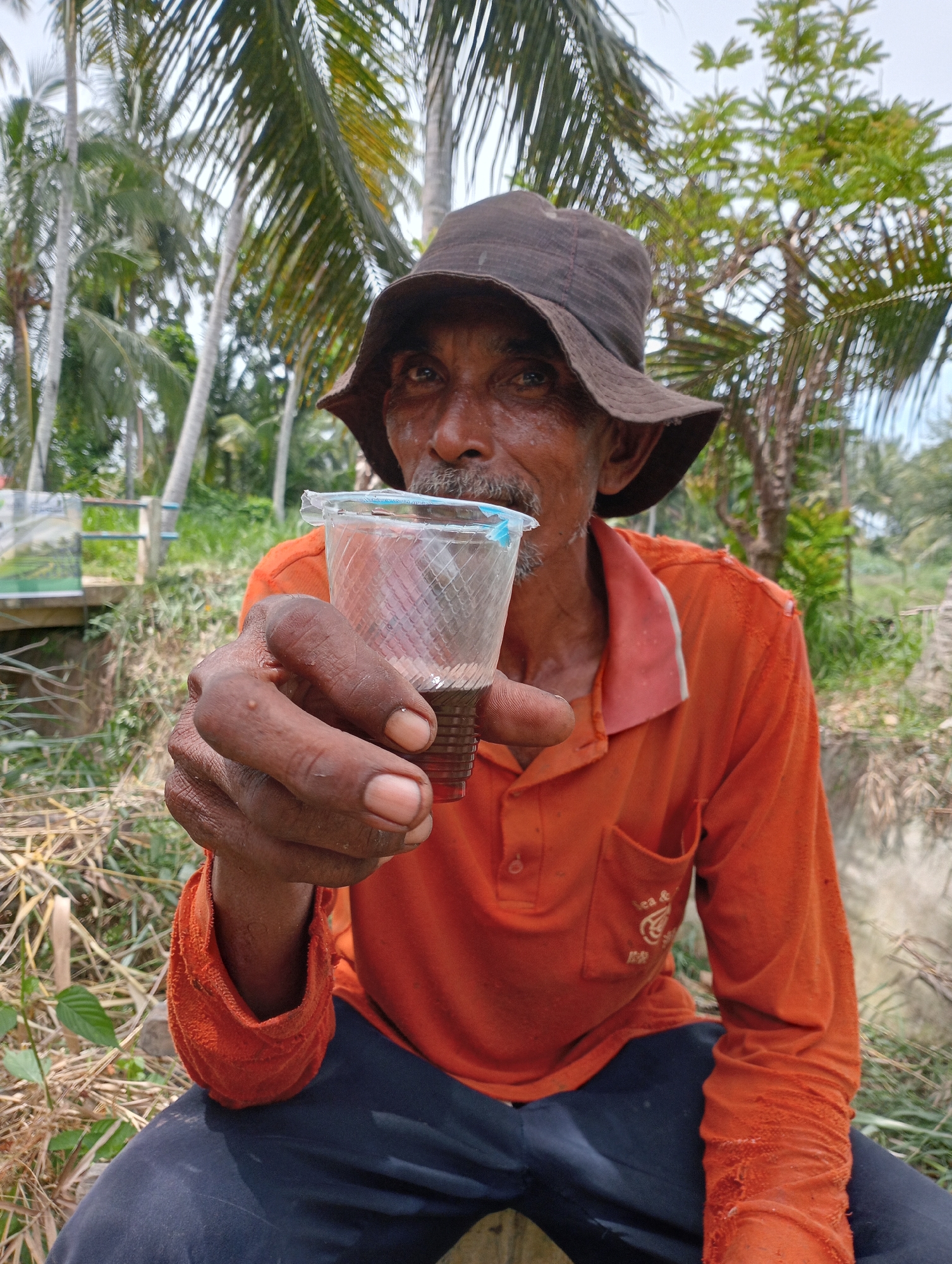 In an outdoor scene, an elderly man with weathered skin and a brown bucket hat is seated, holding up a clear plastic cup filled with a dark liquid. He wears an orange long-sleeved shirt over navy blue pants. The background features tropical vegetation, including palm trees. The location is in the city of Unknown, Unknown.