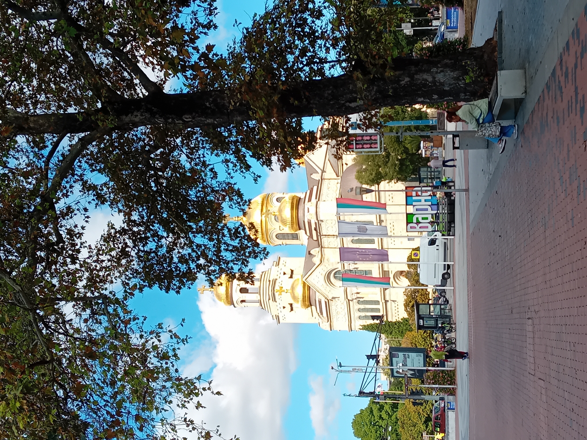The image depicts a wide view of the area around the Cathedral of the Assumption of the Virgin in Varna, Bulgaria. In the foreground, a large tree with autumn foliage partially obscures the view of the cathedral, which stands prominently in the background with its distinctive golden domes. The cathedral is a light-colored stone building with arched windows and features several flags displayed on its facade.
In the midground, there is a paved pedestrian area with a woman sitting on a bench under the large tree, facing away from the camera, with a patterned bag beside her. Across the plaza, there are people walking, bicycles parked, and a white van. A traffic light signal and various signs are visible, including one with the word "Brilliant" and another with "ВАРНА" (Varna). The ground is paved with brick-like tiles.
The environment is urban, with a clear blue sky and scattered white clouds. The overall impression is a daytime scene in a public square.