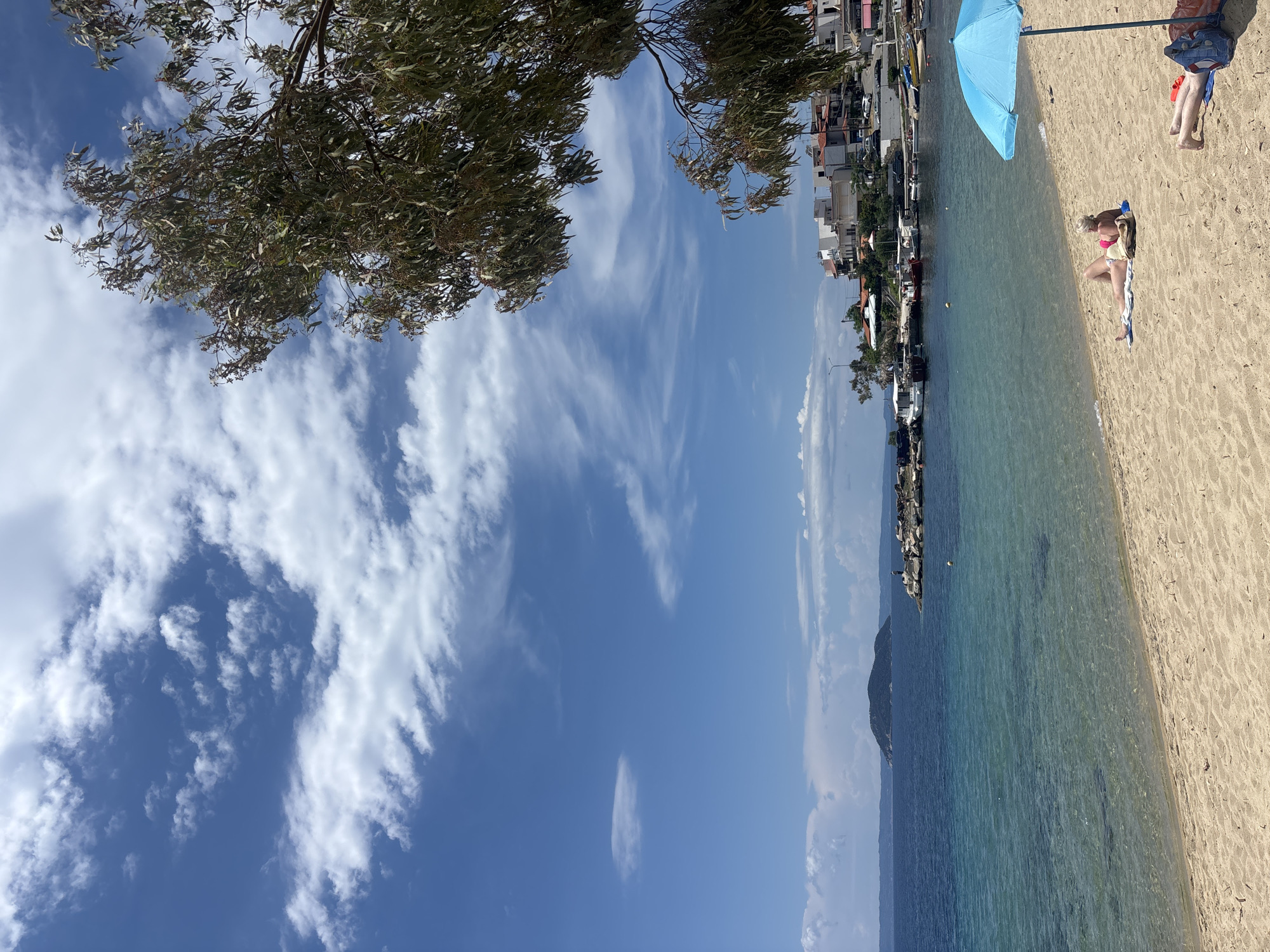 The image captures a beach scene in Sithonia, Greece. The clear turquoise water meets a sandy beach where a woman in a pink bikini lies on a towel, sunbathing. Further along the beach, a light blue umbrella casts a shadow, next to two pairs of legs resting on beach towels. To the left of the beach, a harbor stretches into the distance, lined with docked boats and waterfront buildings. The sky above is a bright blue, scattered with wispy white clouds. A tree with dense foliage occupies the top left corner of the frame, while another tree is on the top right corner of the frame.