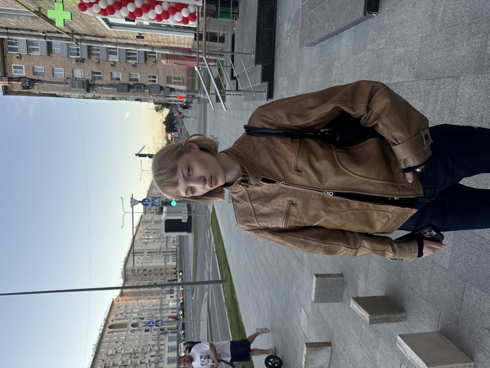 A young woman with blonde, short, choppy hair is posing for a photo on a city sidewalk. She is wearing a brown leather jacket, dark jeans, and a black bag slung over her shoulder. She has one eye closed and her lips pursed. In the background, there is a street with cars, buildings, and pedestrians. A pharmacy with a green cross sign is visible on the right side of the frame, decorated with red and white balloons. The sky is clear with the soft light of dusk.