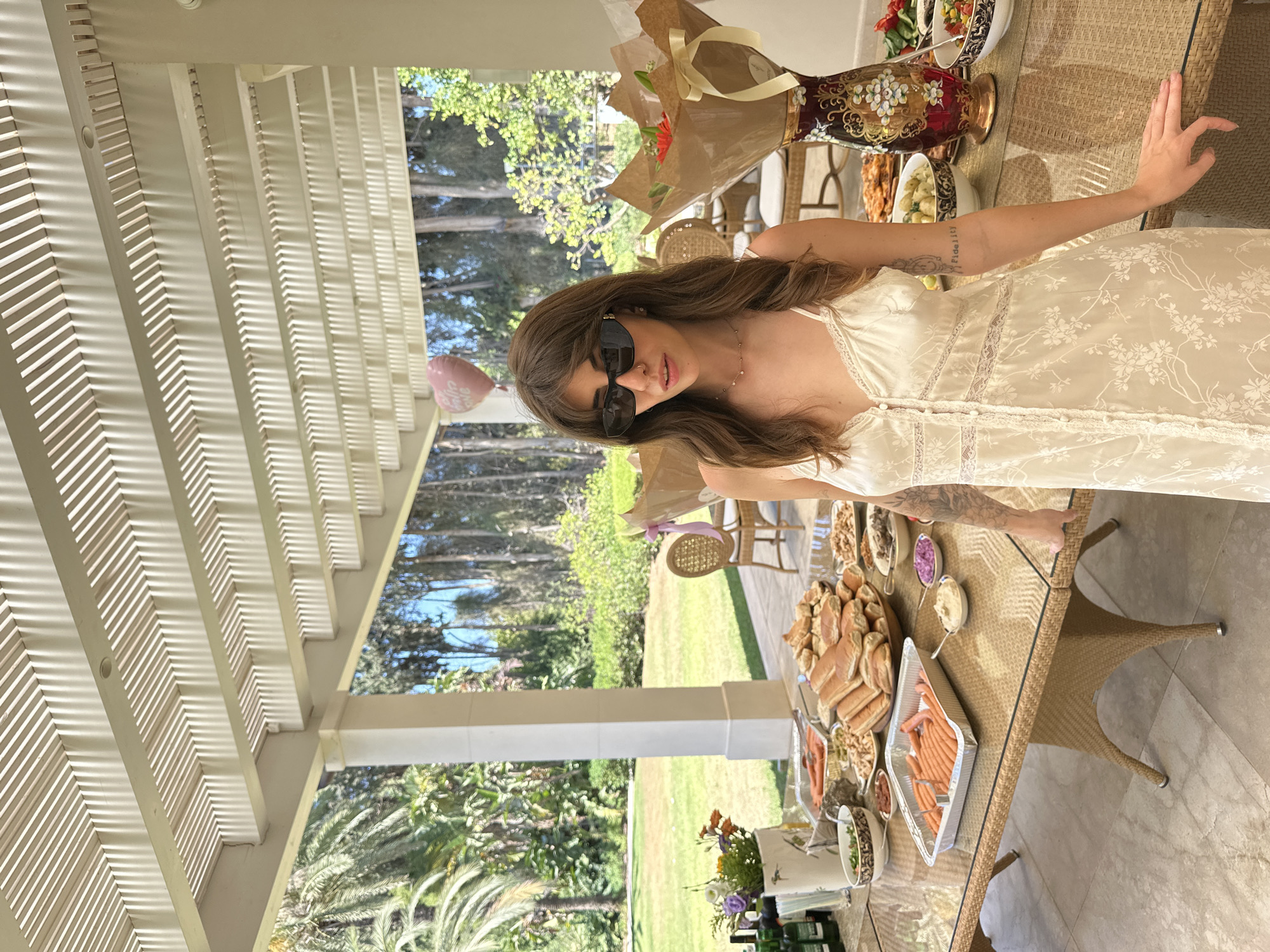 The image depicts a woman standing by a table laden with food and drinks, set up outdoors under a slatted pergola. She is wearing sunglasses, a light-colored dress, and has long brown hair. The table features various dishes, including what appear to be hot dogs, sausages, bread rolls, and salads, along with drinks and floral arrangements. In the background, there is a lush green lawn, palm trees, and other foliage, suggesting a garden or outdoor event space. A pink heart-shaped balloon with text is visible in the upper left portion of the scene. The overall setting suggests a social gathering or party. This scene is located in Savyon, Israel.