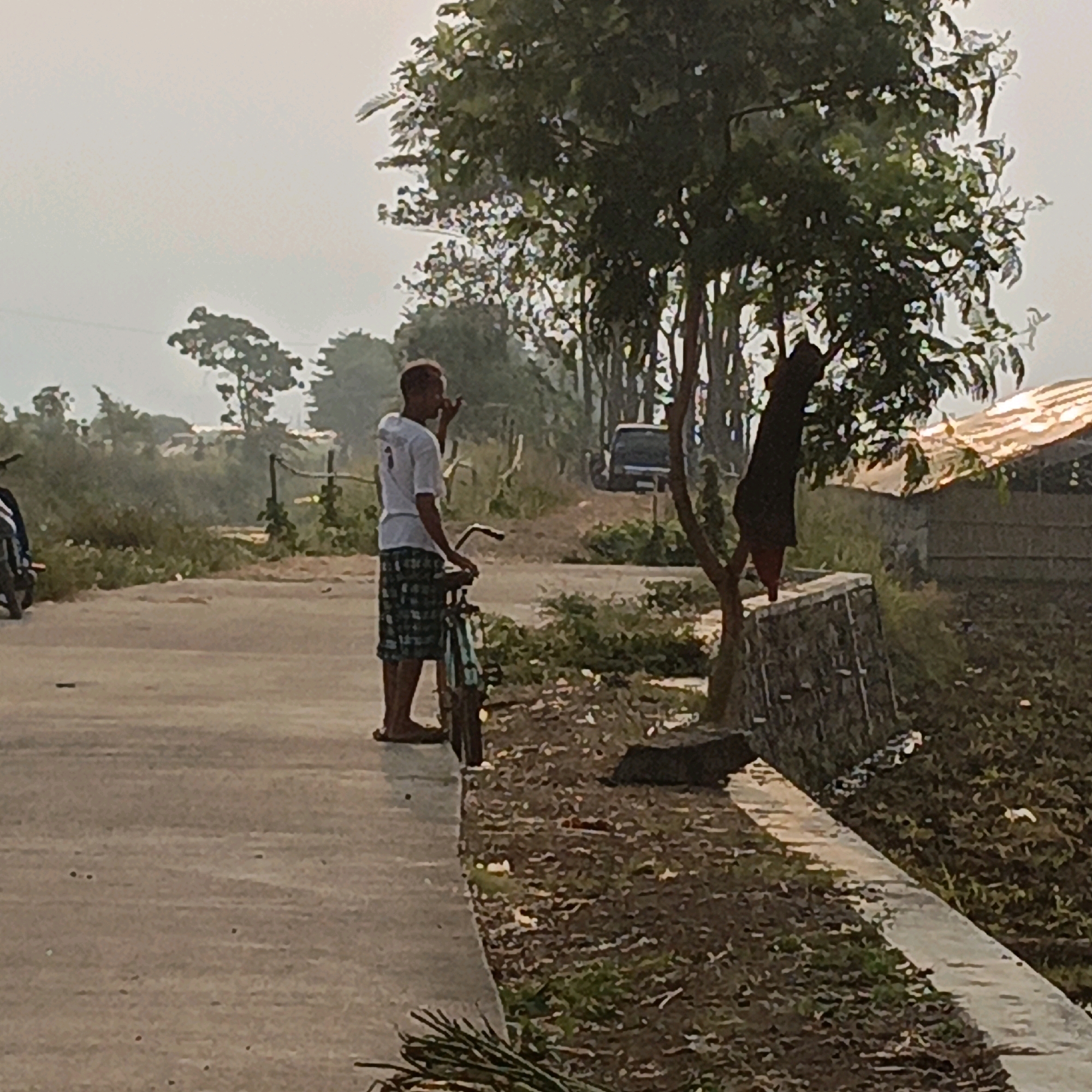 The image depicts a rural outdoor scene in Kesongo, Indonesia, under an overcast sky. In the foreground, a person is standing on a paved road next to a bicycle, holding something to their mouth, possibly smoking. They are wearing a white t-shirt and checkered shorts. Further into the scene, a second person, dressed in dark clothing, is partially visible, appearing to be hanging from a tree branch. The surroundings include vegetation, trees, a stone wall, and a building with a corrugated roof. A vehicle is visible in the background, along with more trees lining a road. The ground appears to be dirt with some patchy grass and debris. The overall impression is that of a quiet, potentially agricultural area.