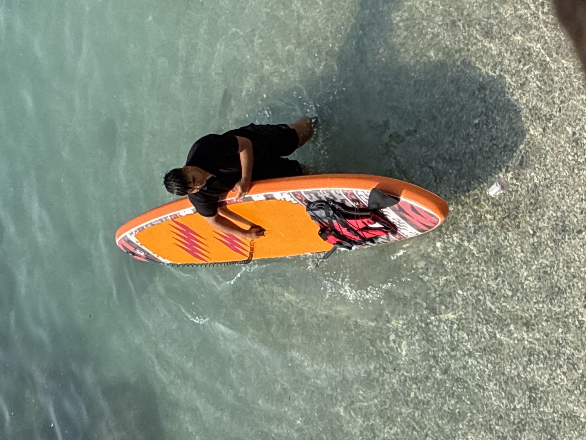 An overhead shot captures a person standing in shallow, clear turquoise water with a paddleboard. The person, wearing a black t-shirt and black shorts, has their hands resting on the paddleboard. The paddleboard is orange with red and white tribal-like markings and a black fin. A red life vest and a black backpack are attached to the paddleboard. The water is clear enough to see the gravelly seabed. A small, clear plastic bottle is floating near the bottom right of the frame. The scene is likely the coastline of Alanya, Turkey, given the characteristic clear waters and seabed.