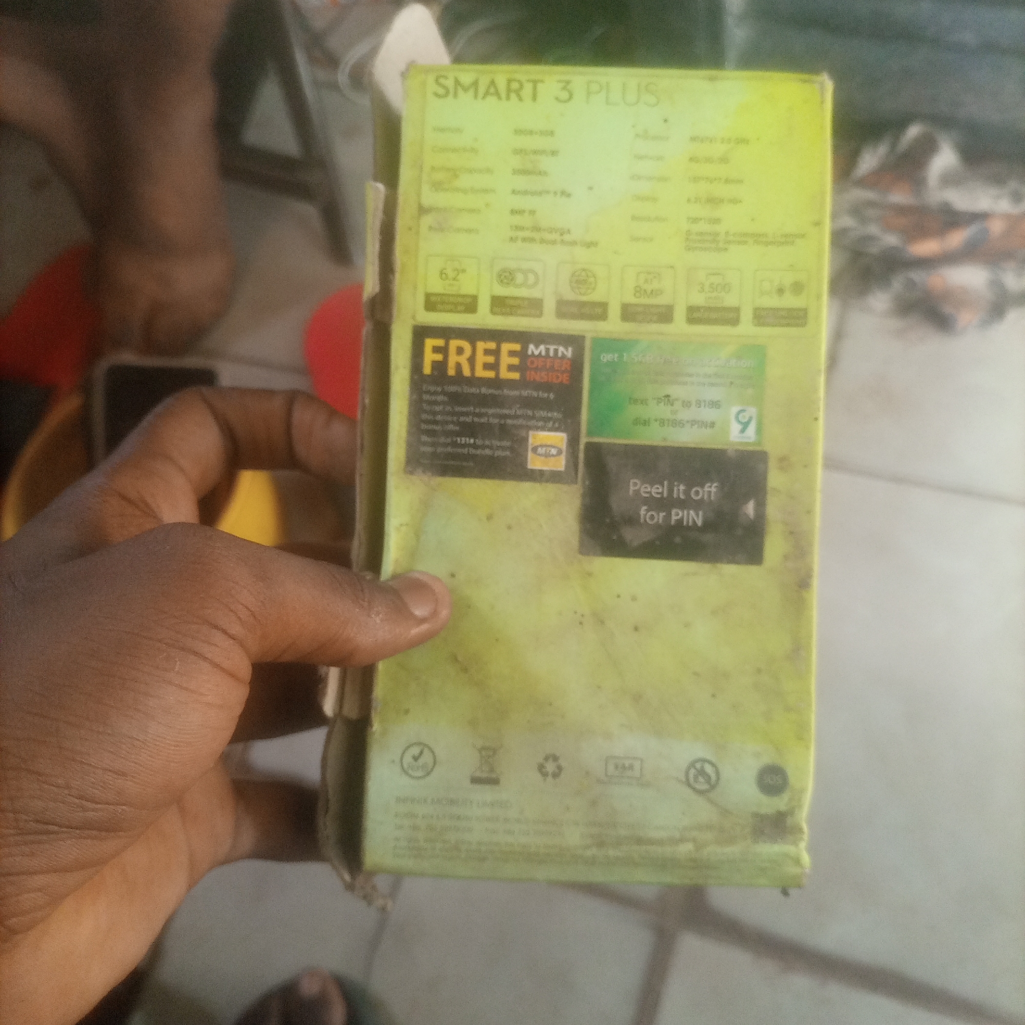 A close-up, eye-level shot captures a hand holding a yellow, rectangular cardboard box, likely for a mobile phone. The box is worn and dirty, with smudges and dark spots scattered across its surface. The box is oriented vertically, with the hand gripping the left side, obscuring some of the box's details.
The top portion of the box displays text that reads "SMART 3 PLUS". Below this, a grid of specifications is visible, including camera resolutions (e.g., "8MP", "6.2\"") and battery capacity (e.g., "3,500 mAh"). A prominent "FREE" banner with "MTN OFFER INSIDE" is situated on the lower left of the box, along with instructions for redeeming a data bonus. On the right side, a green section encourages the user to "get 1.5GB data", and instructs them to "Peel it off for PIN".
The background is blurred and out of focus, showing a dimly lit indoor environment. To the left of the box, a person's lower legs and feet are partially visible, suggesting they are sitting or lying down. The overall lighting is dim, creating a muted color palette dominated by the yellow of the box and the muted tones of the background. The scene appears to be an indoor setting, possibly a home or a shop.