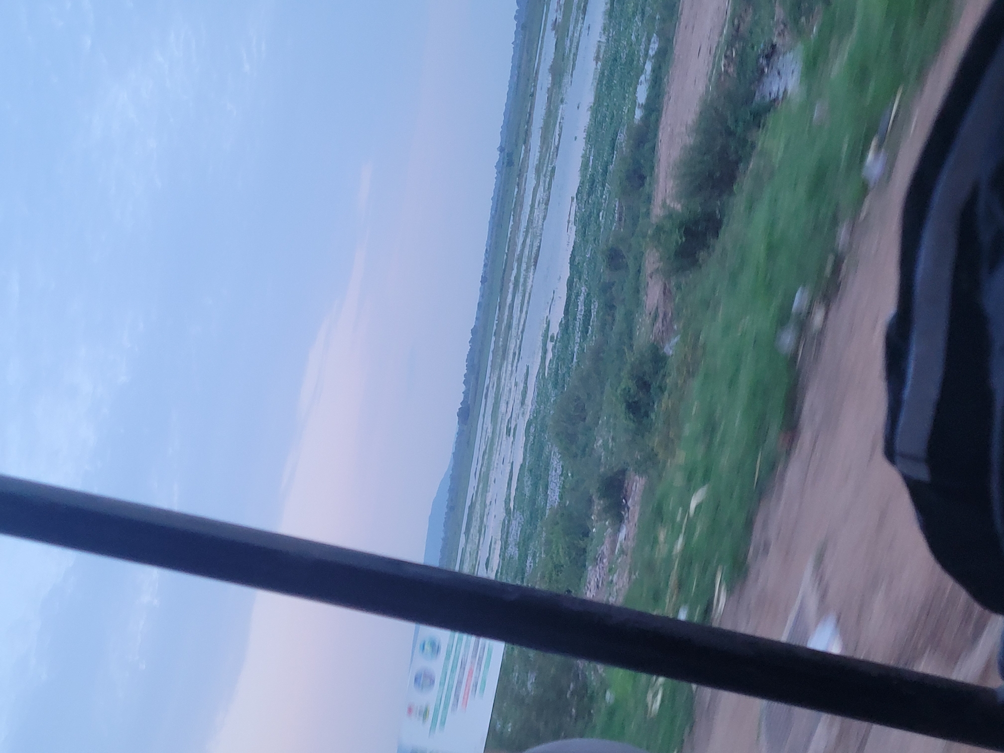 This media file captures a scenic view of a wide, shallow body of water, likely a river or floodplain, surrounded by lush green vegetation and a dirt path. The water is interspersed with patches of greenery, suggesting it may be a wetland or a flooded agricultural area. In the background, a faint silhouette of hills or mountains can be seen against a pale blue sky. The sky itself is partly cloudy, with wispy clouds scattered across it.
A dark, thick horizontal bar, possibly a railing or a part of a vehicle, traverses the foreground, obscuring a portion of the scene. To the right, a dark, blurry object, possibly the edge of a vehicle or a person wearing dark clothing, is partially visible.
A small section of a white sign with some text and graphics is visible at the bottom left of the image. The text is mostly illegible due to the angle and blurriness, but some colorful graphics and what appear to be circular icons are discernible.
The overall impression is one of a serene, natural landscape, possibly captured during an early morning or late afternoon due to the soft, diffused light. The scene suggests a rural or semi-rural setting in Lokoja, Nigeria, a region known for its confluence of major rivers and expansive natural beauty. There are no clear indications of specific activities or events, beyond the implied movement of the observer capturing the scene, likely from a moving vehicle or a vantage point.