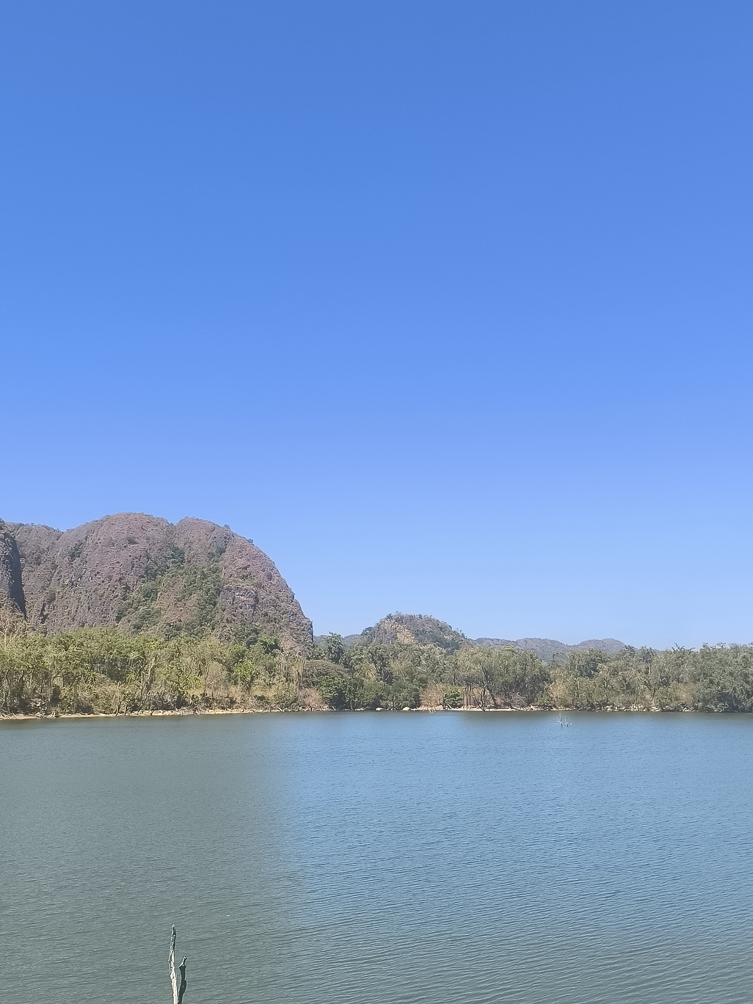A landscape image shows a large body of calm water in the foreground, with a dense line of trees and vegetation along the far bank. Beyond the trees, rugged, rocky hills rise up under a clear blue sky. The terrain appears arid and mountainous. The scene is devoid of any human presence or man-made structures, suggesting a natural and undeveloped environment. The location is identified as the city of Kalabbirang, Indonesia.