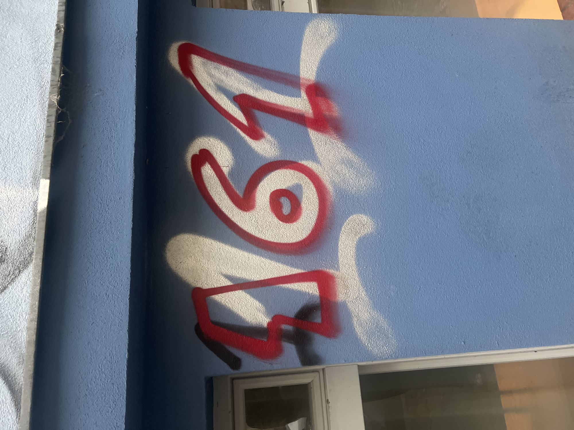 The image shows a close-up, eye-level, outdoor shot of graffiti spray-painted in red and white on a textured blue wall. The graffiti spells out the numbers "161" in a stylized, graffiti font. A white outline and shadow effect is applied to the numbers. To the left of the graffiti, a white-framed window with a glass pane is visible. The wall appears to be part of a building exterior. The image does not provide enough information to determine the specific location within Berlin, Germany, or to identify any people or specific actions.