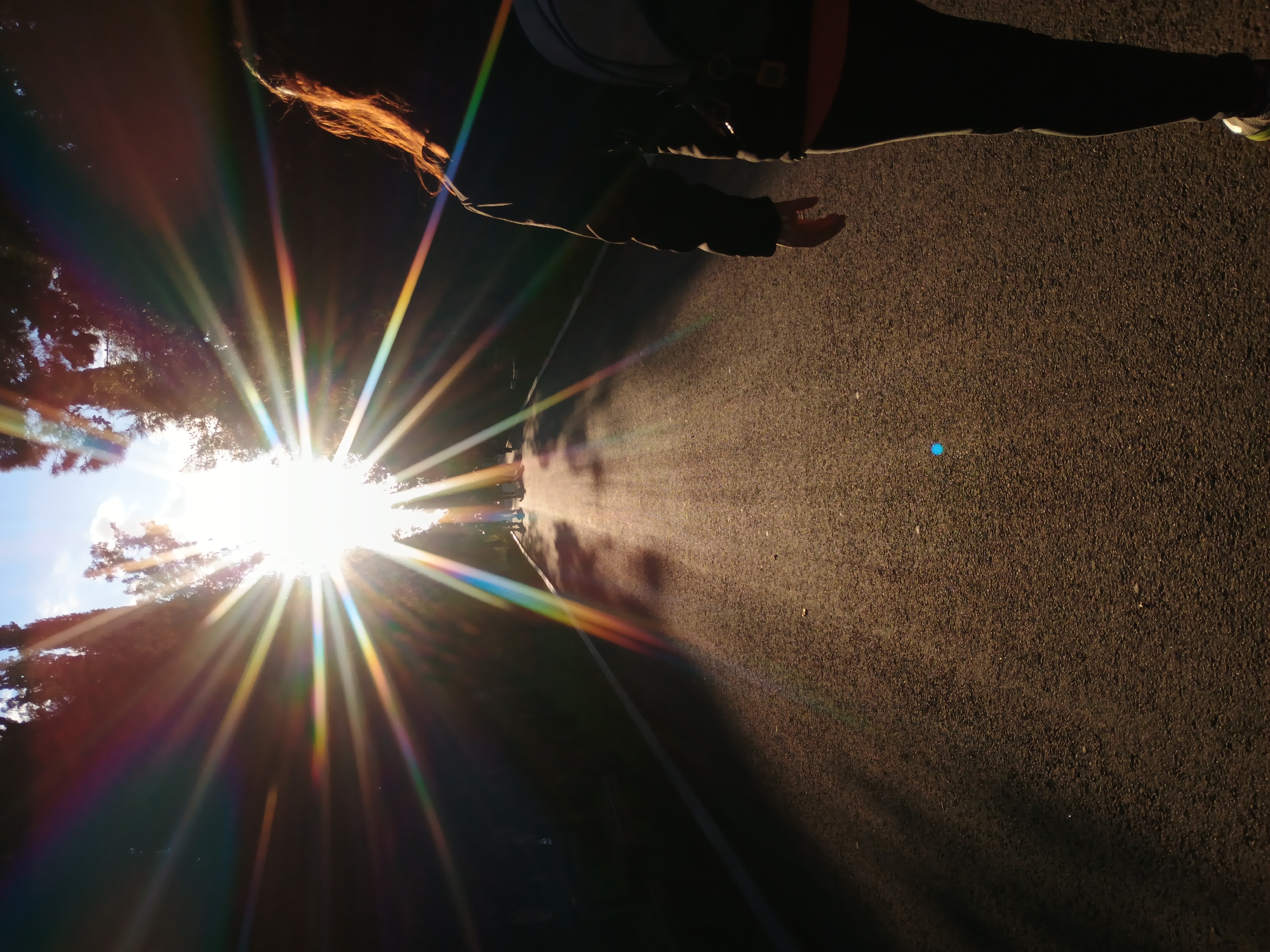 The image shows a person walking on an asphalt road. The sun is shining brightly through trees in the background, causing a starburst effect with rainbow flares. The person is wearing dark clothing and the legs of the person are partially visible in the upper right of the image, with part of one foot visible. Part of the person's hair is red and visible. The road surface is textured and dark, with patches of brighter light. The city of Stara Zagora, Bulgaria is not discernible in the image, nor is any identifiable location, though the presence of trees and an asphalt road suggests an outdoor public area.
