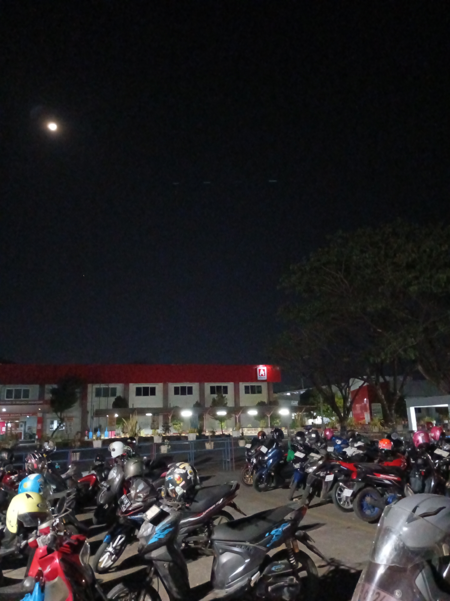 This is a nighttime photograph taken in a city, possibly Makassar, Indonesia, showing a parking lot filled with motorcycles. A large, dark building with a red roof and white windows is visible in the background, featuring the red letter "A" logo. A large tree with dark foliage is situated to the right of the building. The sky is dark, with a bright moon visible in the upper left quadrant of the image. Several motorcycles are parked closely together in the foreground, with various helmets placed on top of them. The lighting is dim, suggesting it is late at night.