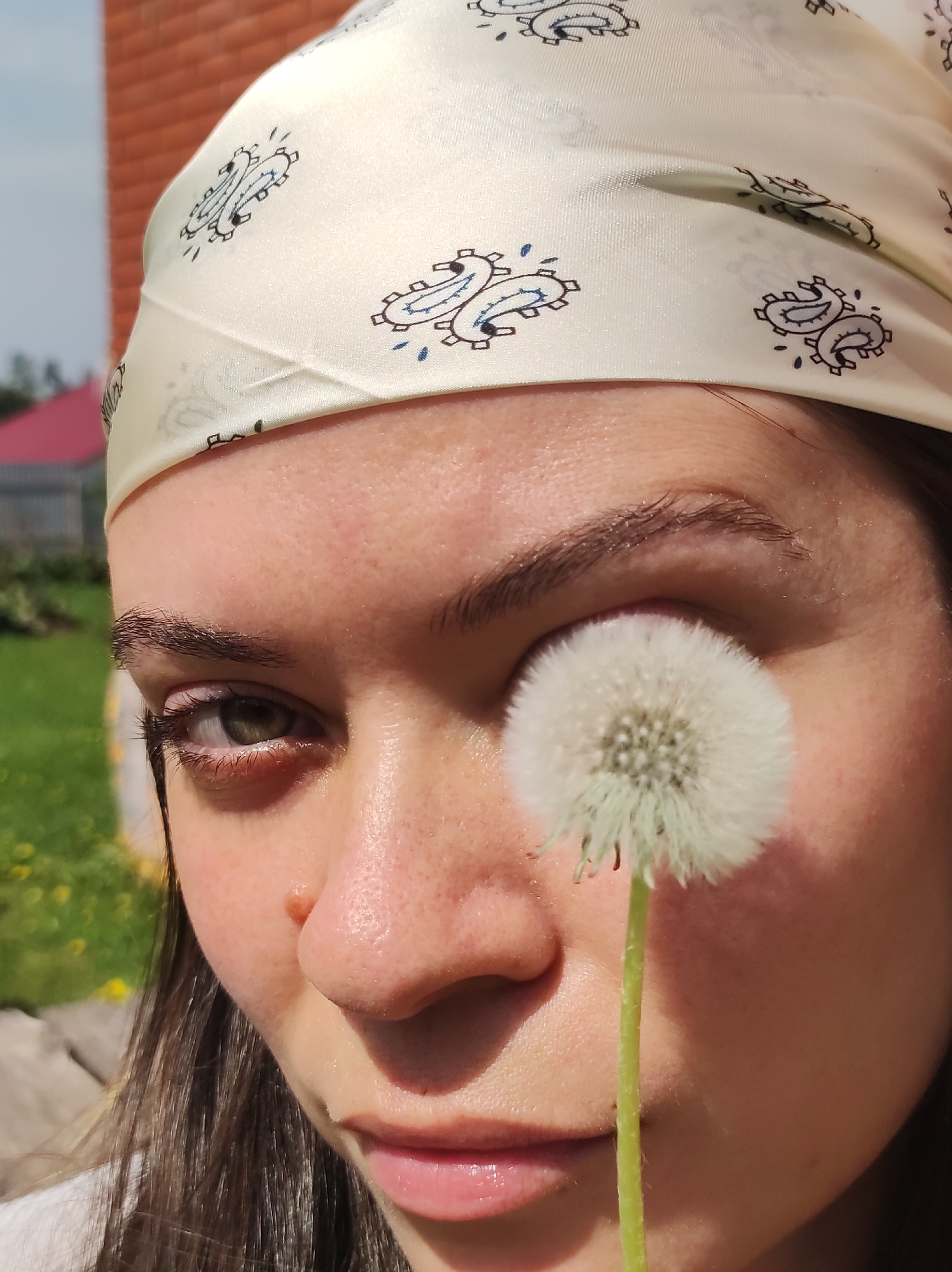 A close-up of a person's face. The person wears a white headscarf with a paisley-like pattern. Their right eye is obscured by a dandelion seed head. The background includes a brick structure and green vegetation, suggesting an outdoor location in Фоки, Russia.