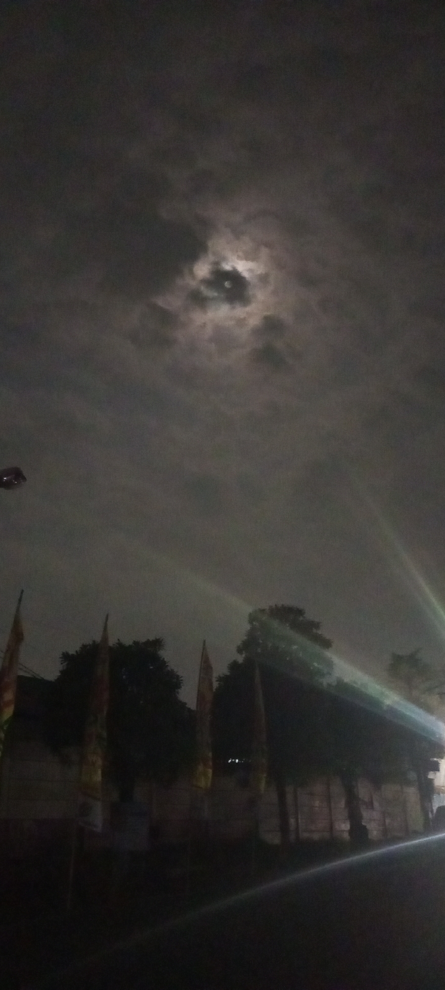 This vertical, low-light photograph captures a night scene in the city of Setu, Indonesia. The sky is predominantly dark and cloudy, with the moon visible and partially obscured by clouds, creating a dramatic effect. Streaks of light, likely from a nearby light source, cut diagonally across the frame from the upper right. In the foreground, silhouetted against the sky, are trees and several flags on poles. The flags appear to have some design or pattern, but are not clearly discernible due to the darkness. Below the trees and flags, a dark, indistinct structure, possibly a building or fence, runs horizontally across the lower portion of the image. The overall impression is one of a quiet, possibly rural or suburban, night in Indonesia.