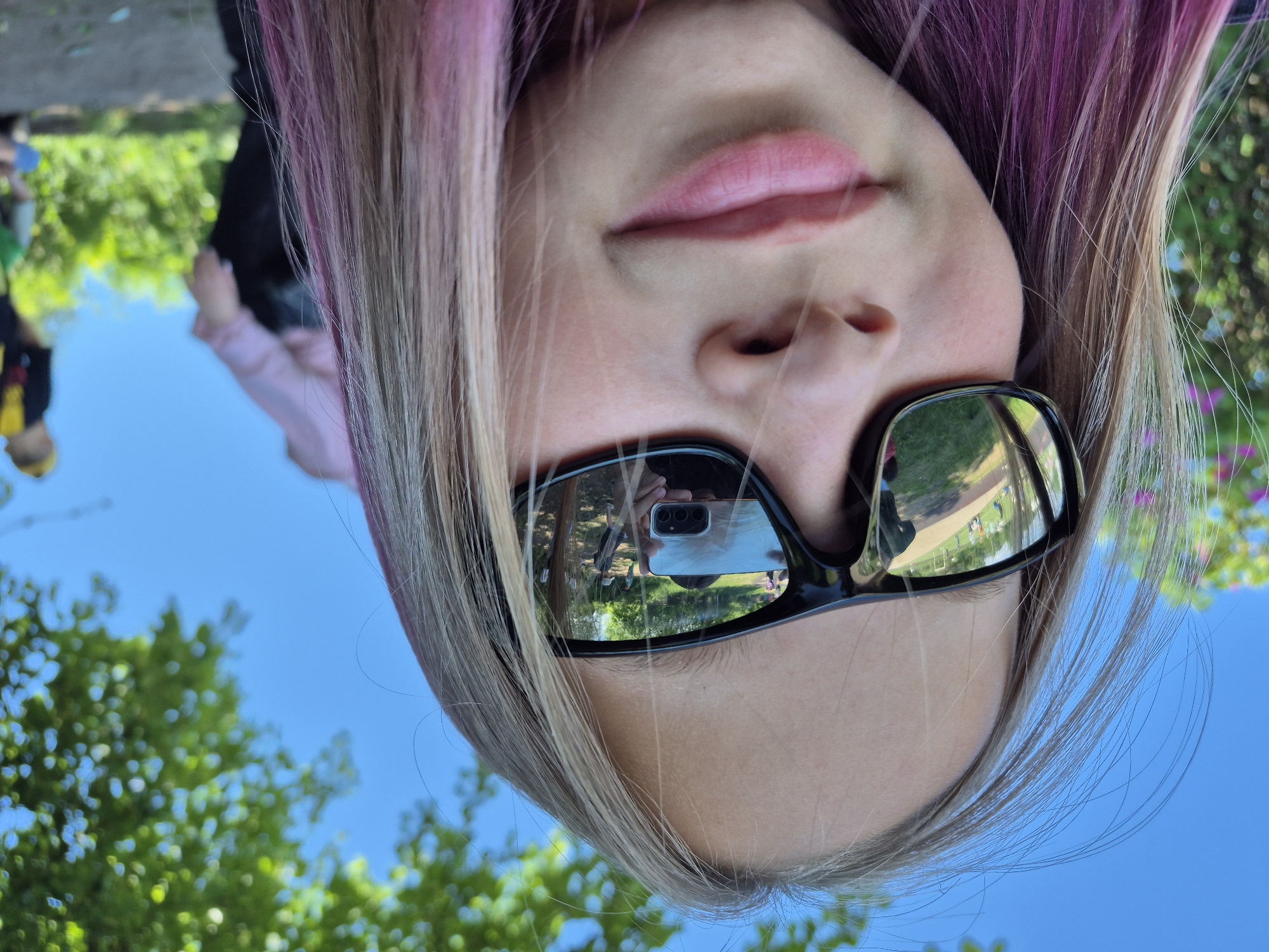 A close-up, wide-angle selfie shows a young person wearing black sunglasses, with their face filling most of the frame. The person has fair skin, light brown hair with pink streaks, and is smiling slightly. The sunglasses reflect a scene of other people and a phone being used to take the photo. In the background, out of focus, are trees and a bright blue sky. The overall setting appears to be outdoors, possibly a park or recreational area. No specific landmarks are visible to determine a location in Chaikovsky, Russia.
