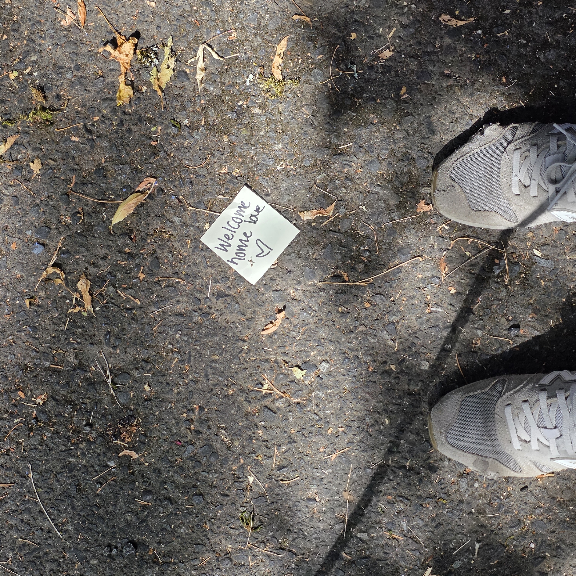 A pair of gray sneakers are positioned on asphalt, which is littered with small stones and fallen leaves. In the center of the frame, a white note is placed on the ground. The note is written in black marker and says "Welcome home love" with a heart symbol below the text. The scene appears to be outdoors, possibly on a sidewalk or driveway, with shadows indicating sunlight filtering through trees. The location is Portland, United States of America.