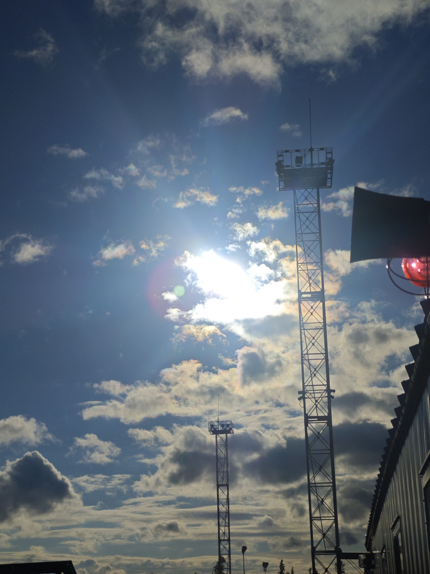 The image captures a daytime scene with a bright sun partially obscured by clouds in a blue sky. Two metal lattice communication towers are visible, one significantly taller than the other. The taller tower is positioned to the right, with a platform at the top. To the right of the taller tower, part of a building's corrugated metal roof and wall are visible, along with a red light fixture and what appears to be a speaker. The smaller tower stands farther away to the left. The foreground includes obscured structures that could be part of buildings or other installations. The overall environment suggests an industrial or infrastructure-related area. The location is Unknown, Unknown.