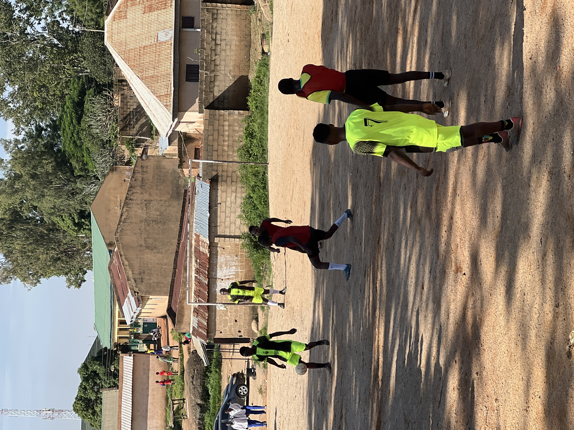 The image depicts a group of young men playing soccer on a dirt field in Jos, Nigeria. The scene is characterized by a dusty, sun-drenched environment with buildings in the background. The players are dressed in athletic attire, with some wearing brightly colored jerseys. One player in the foreground is actively kicking a soccer ball, while others are positioned around the field, engaged in the game. The buildings in the background appear to be a mix of residential and possibly educational structures, with corrugated iron roofs visible. The overall impression is one of casual sport and community activity within an urban African setting.
