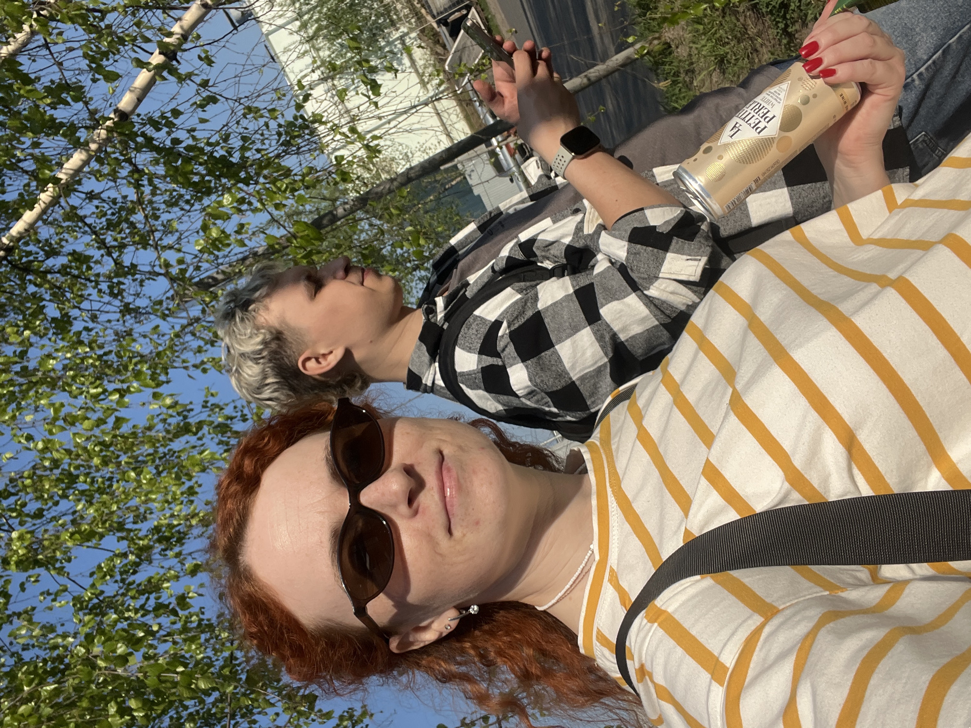The image presents an outdoor scene with two individuals. One person, positioned closer to the camera, has red hair and wears sunglasses, a white and yellow striped shirt, and a necklace. The second person, situated behind, has short, bleached hair and is wearing a black and white plaid shirt. They appear to be holding a smartphone and a can of La Petit Perle White Wine. They seem to be situated outdoors, possibly in a park, based on the surrounding greenery and birch trees with new leaves. In the background, there is a glimpse of what appears to be buildings or structures in an urban setting. This location is near the city of Chaikovsky, Russia.