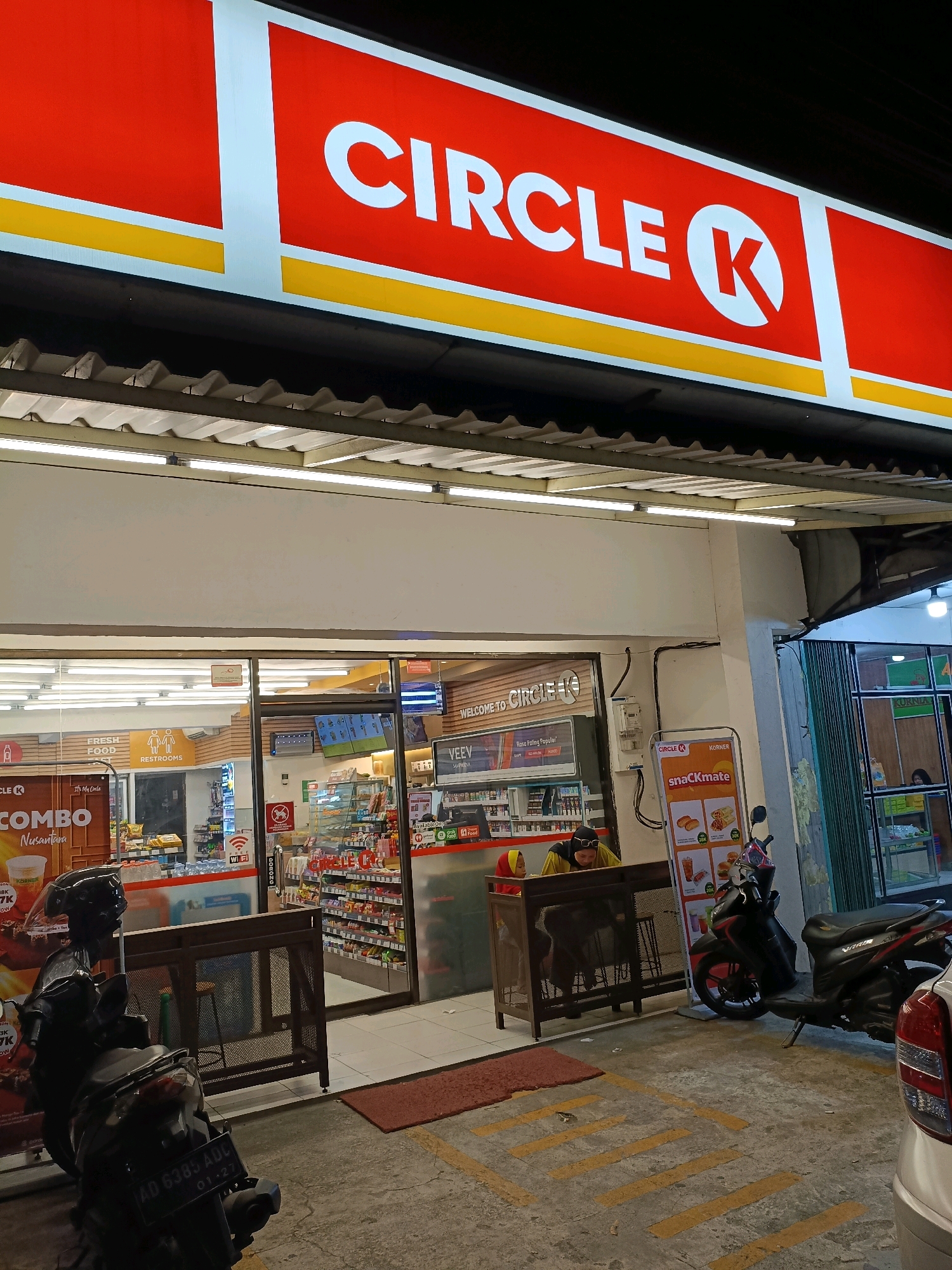 The image captures the exterior of a Circle K convenience store in Purwomartani, Indonesia, at night. The prominent red and white Circle K sign is illuminated, displaying the brand's logo. Below the sign, a white overhang shelters the entrance. The store's glass doors reveal shelves stocked with merchandise, and fluorescent lights illuminate the interior. In front of the store, two individuals, likely employees, are visible behind a counter, one wearing a yellow shirt and headscarf, and the other a red headscarf. Outside the store, a dark-colored scooter is parked, with its license plate readable as "AD 6385 ADC 01-27". Further to the right, a light-colored car is partially visible. The ground outside the store is paved with concrete, marked with yellow lines, and a red mat is placed near the entrance. The overall environment suggests a typical convenience store scene in a public area.