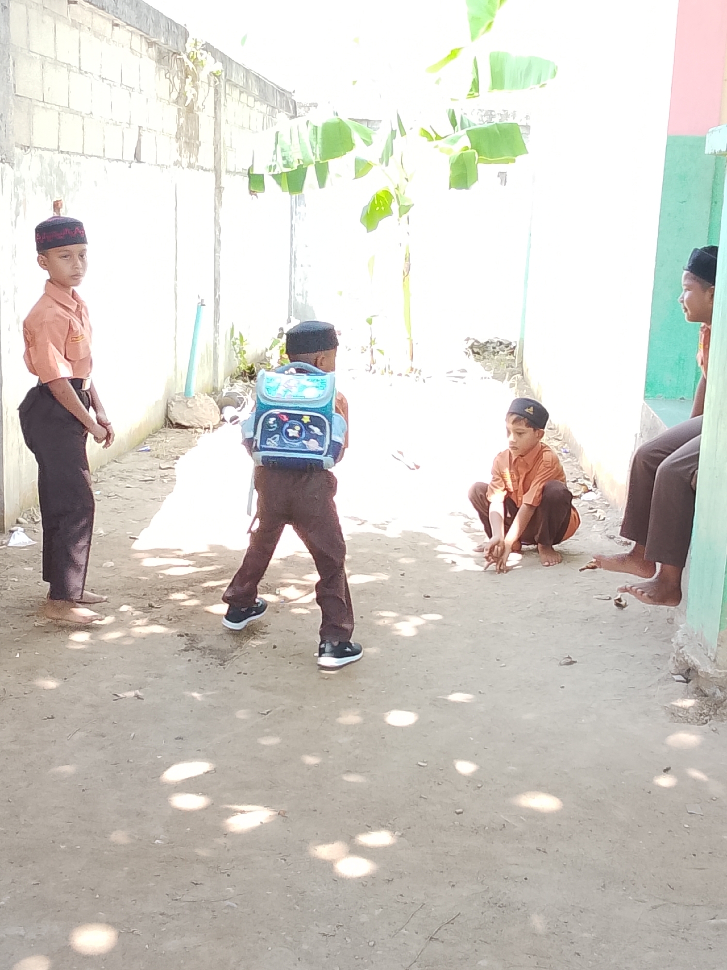 This image shows four young boys in an outdoor area, likely in Baratpayaitek, Indonesia. The ground is dirt and sandy, with dappled sunlight filtering through unseen foliage. In the background, there is a light-colored brick wall and some large green leaves, suggesting a tropical or subtropical environment.
On the left, a boy wearing a tan shirt, dark pants, and a patterned red and black cap stands facing the camera. He has his hands clasped in front of him. Next to him, to the right and slightly further back, is another boy facing away from the camera, wearing a tan shirt, dark pants, and a black cap. He is carrying a blue backpack with cartoon characters on it.
In the middle of the frame, a boy in a tan shirt, dark pants, and a black cap is crouched down, facing left. He appears to be playing a game with small objects on the ground. To the right of the crouching boy, another boy in a tan shirt, dark pants, and a black cap is seated against a wall. His legs are extended, and he is looking towards the left. A portion of a green wall is visible behind him.